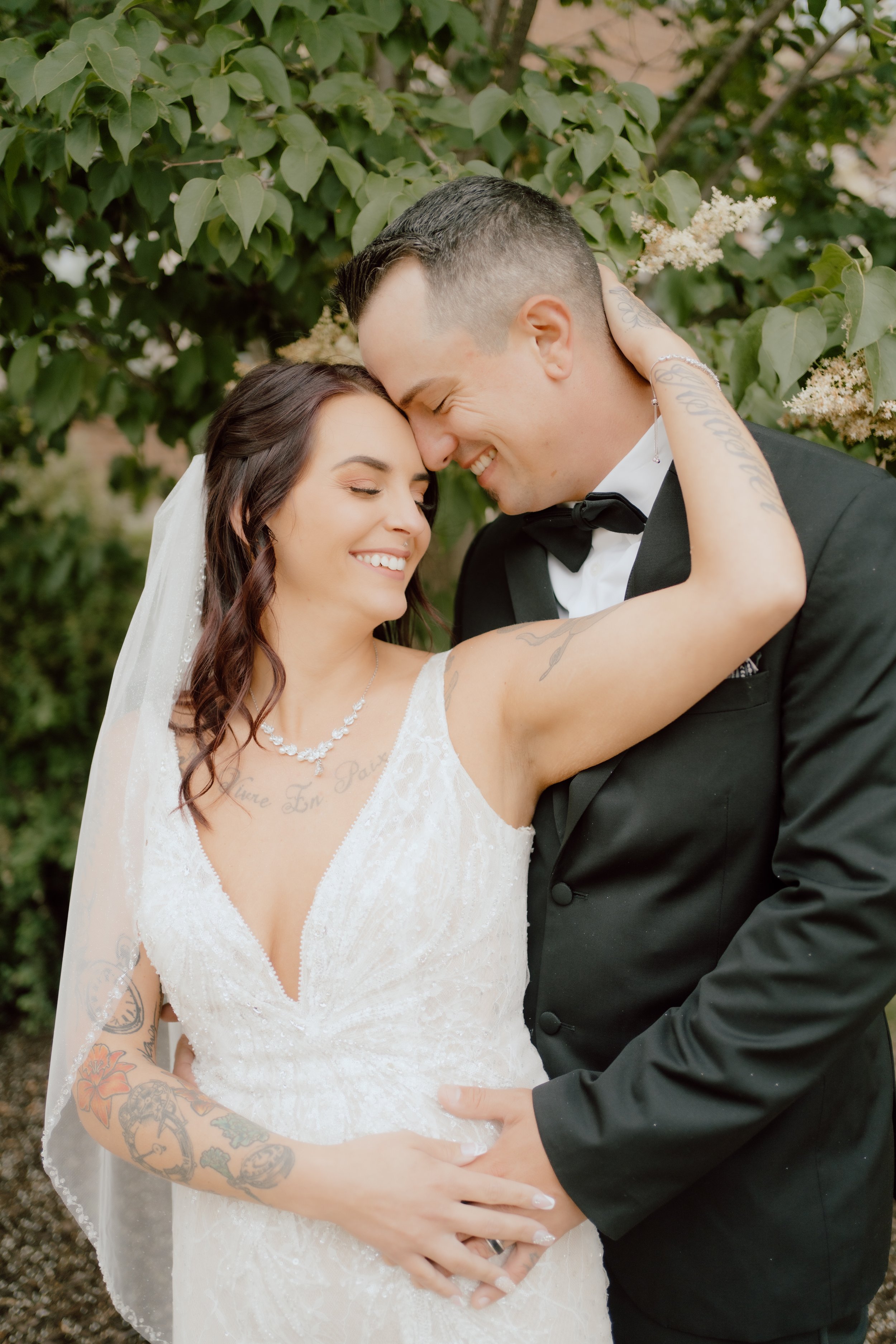 A bride and groom share a close, joyful moment with their foreheads touching and eyes closed, outdoors with leafy green background, the bride wearing a white lace wedding dress and the groom in a black tuxedo.
