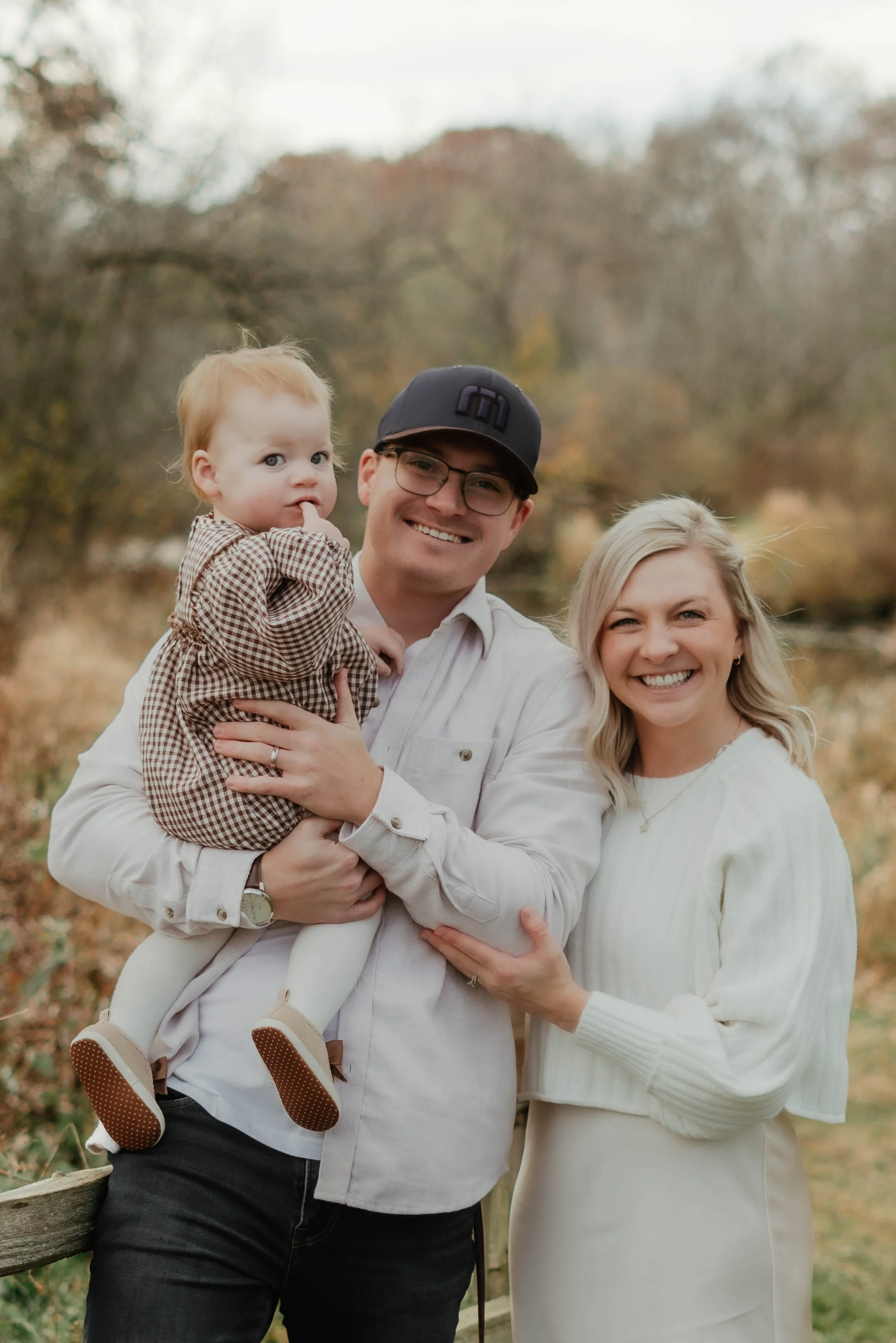 A smiling family of three outdoors in autumn: a man holding a young girl, and a woman standing next to them, all dressed in neutral tones with fall foliage in the background.