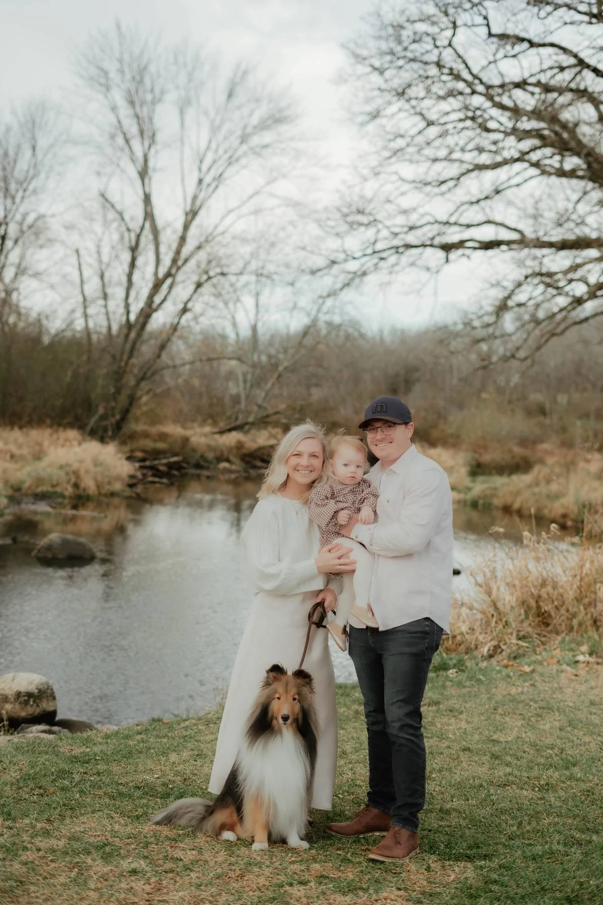 A family of three with their dog standing outdoors near a creek, with leafless trees and a cloudy sky in the background during fall or winter.
