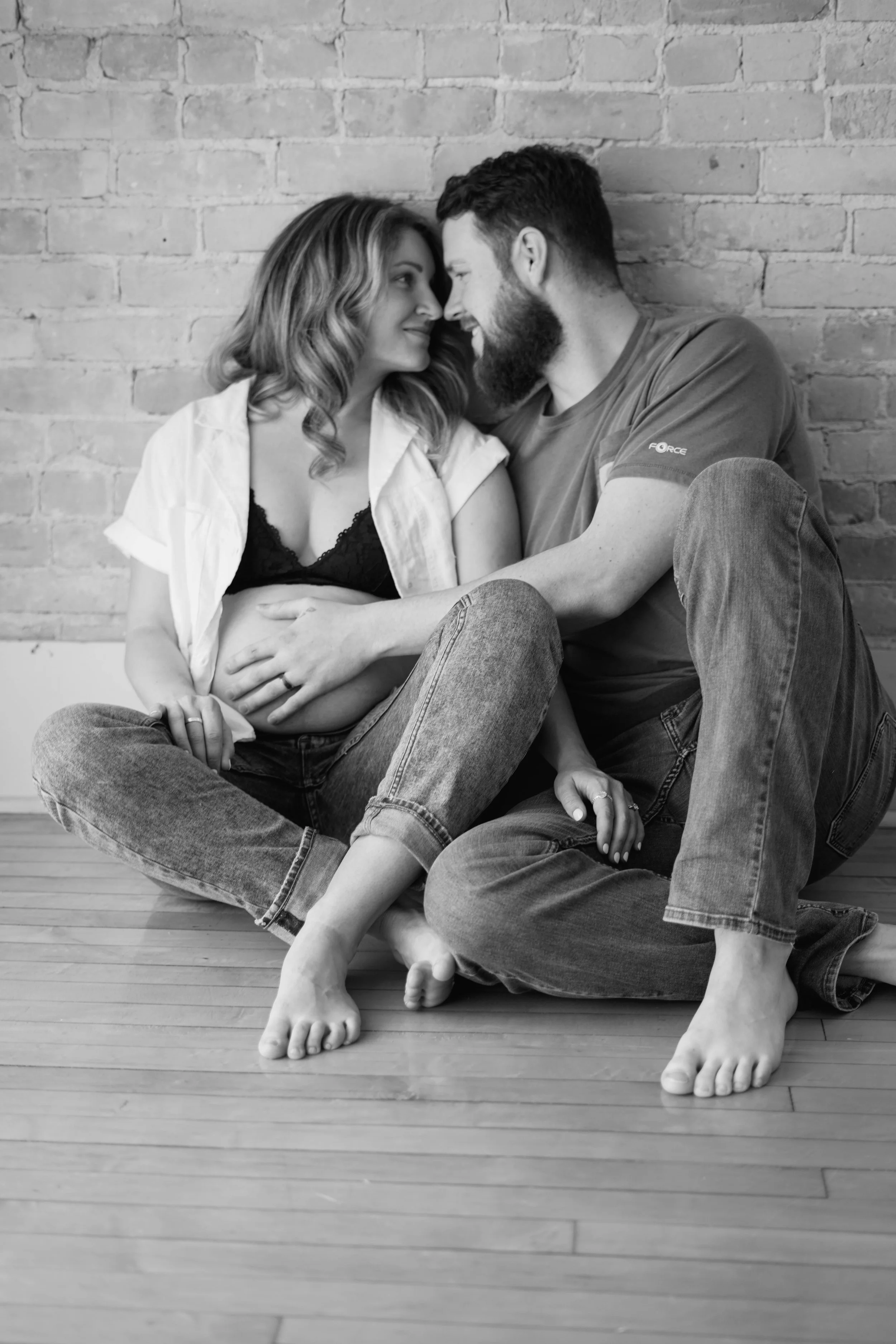 A black and white photo of a couple sitting on the floor against a brick wall, touching foreheads and smiling at each other, with the woman showing her pregnant belly.