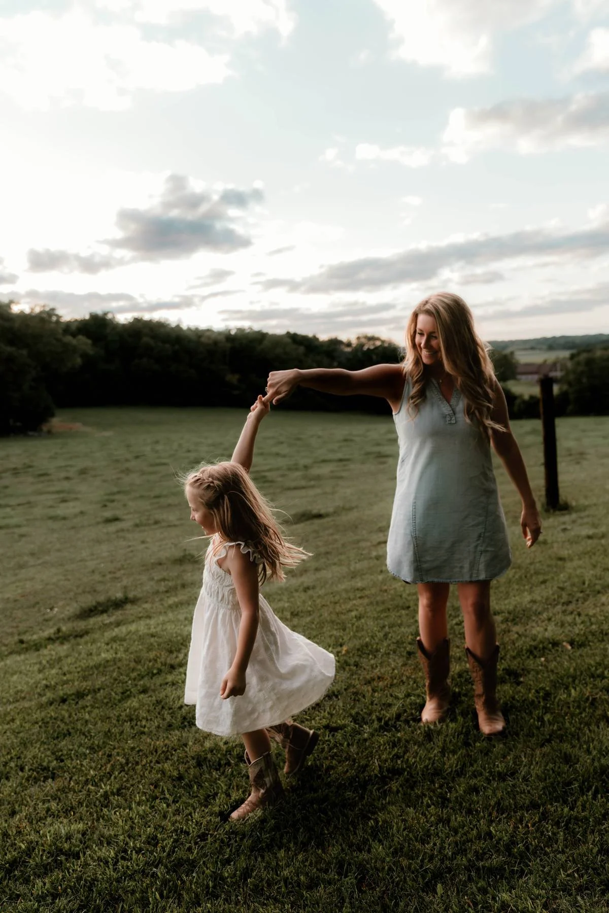 A woman and a young girl dancing in a grassy field during sunset, holding hands and smiling.