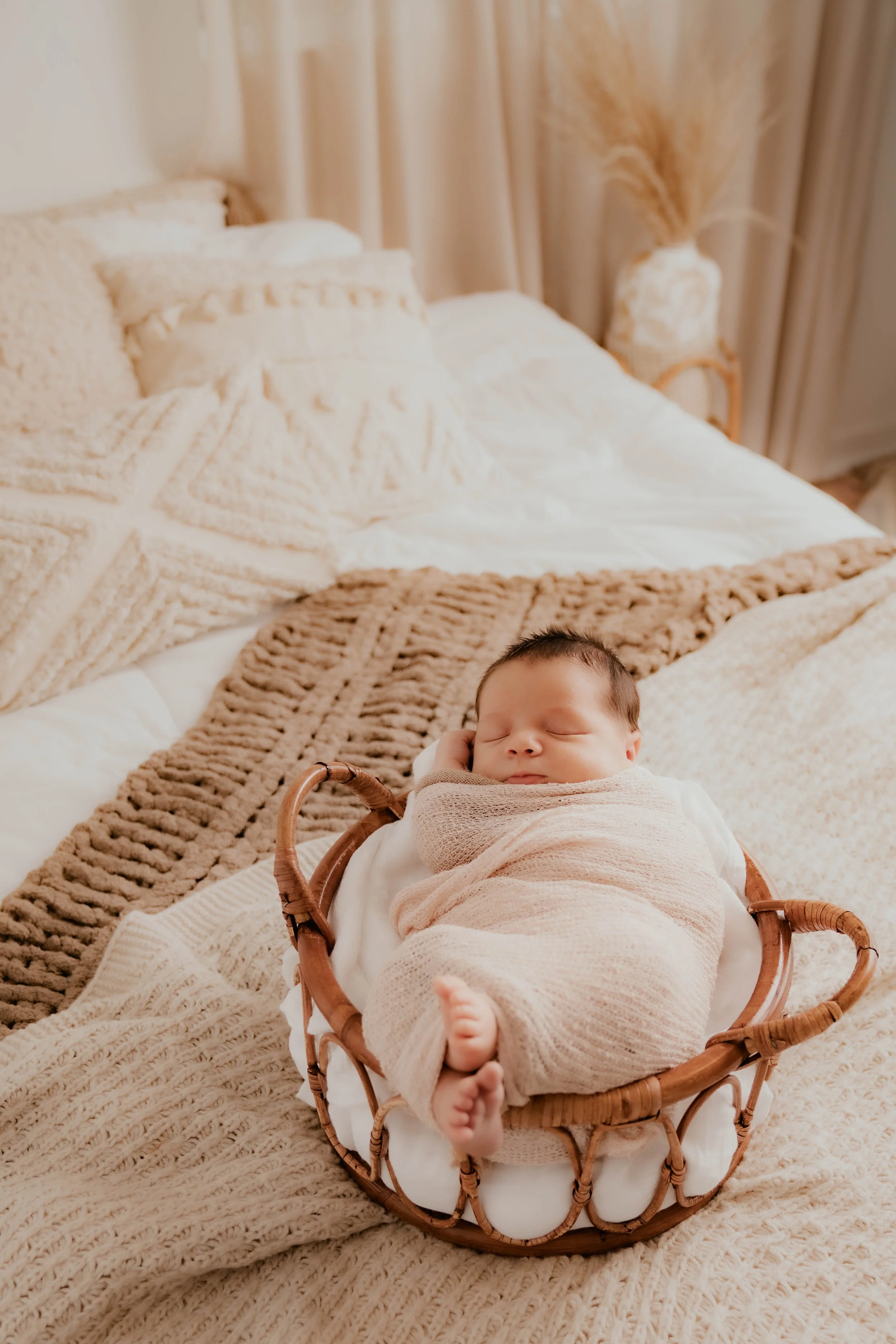A sleeping baby wrapped in a pink blanket inside a small wicker basket on a bed with beige and cream-colored bedding.