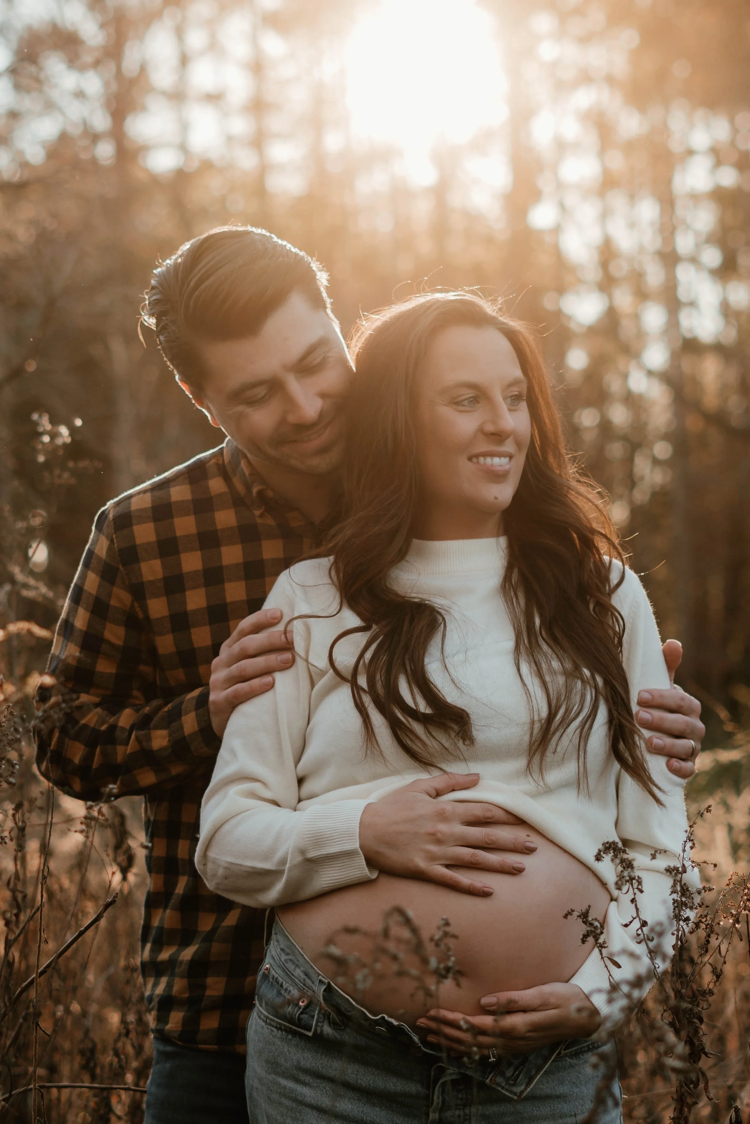 A pregnant woman with long brown hair smiling as a man, likely her partner, stands behind her with his arms around her shoulders in a warm outdoor setting during sunset.