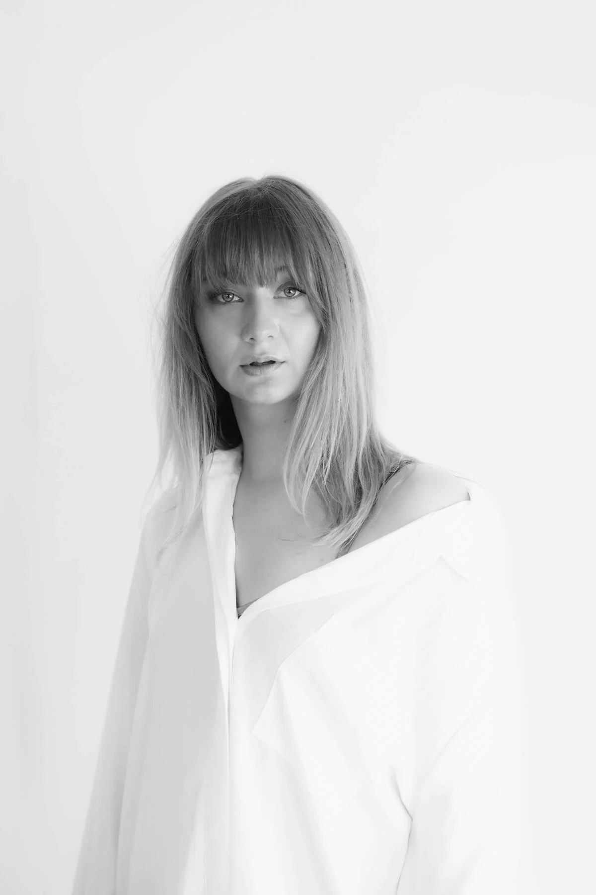 A black-and-white portrait of a woman with long hair and bangs, wearing a loose white shirt, against a plain light background.