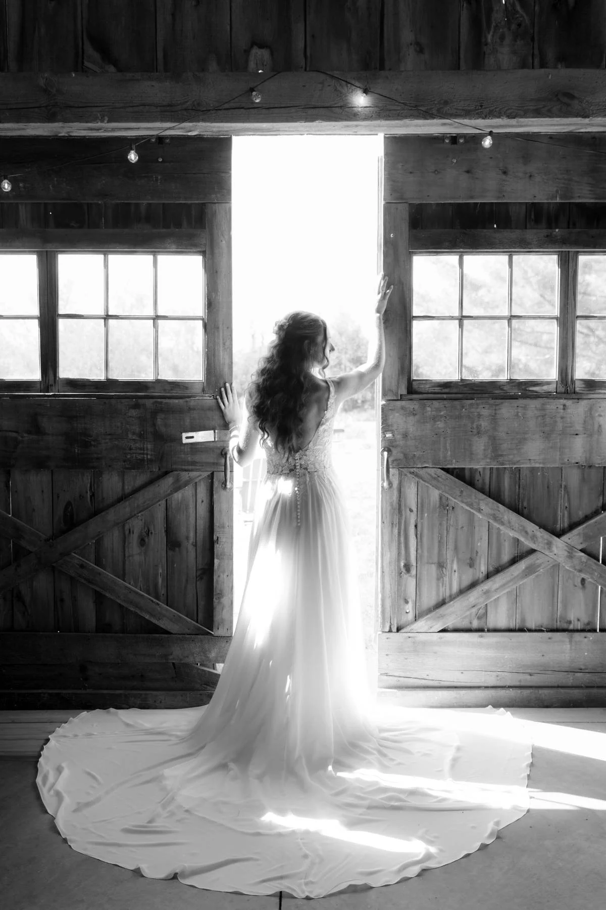 A woman in a wedding dress standing in front of open barn doors, looking outside with light streaming in.