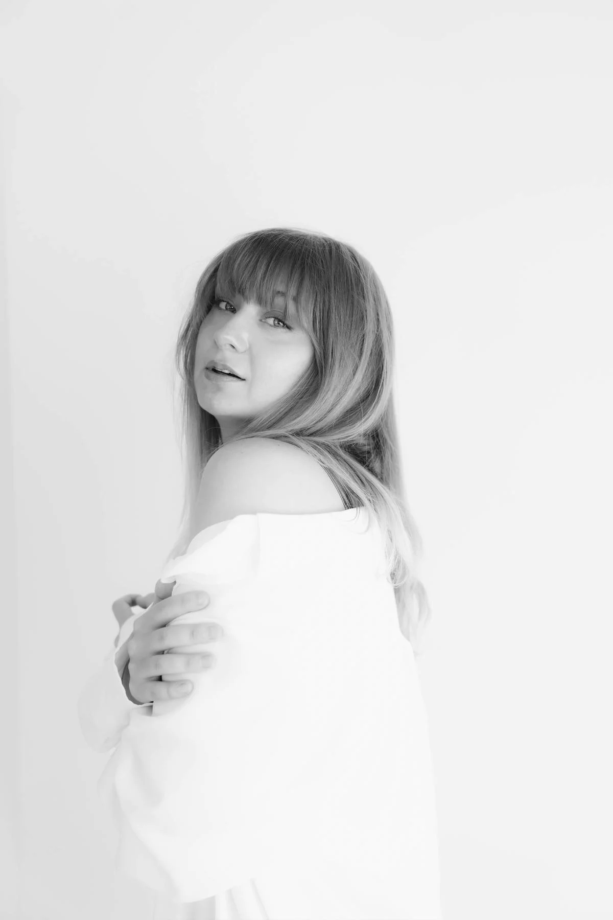 Black and white photograph of a woman with long hair, looking at the camera with a gentle expression, wrapped in a white cloth or blanket, standing against a plain background.
