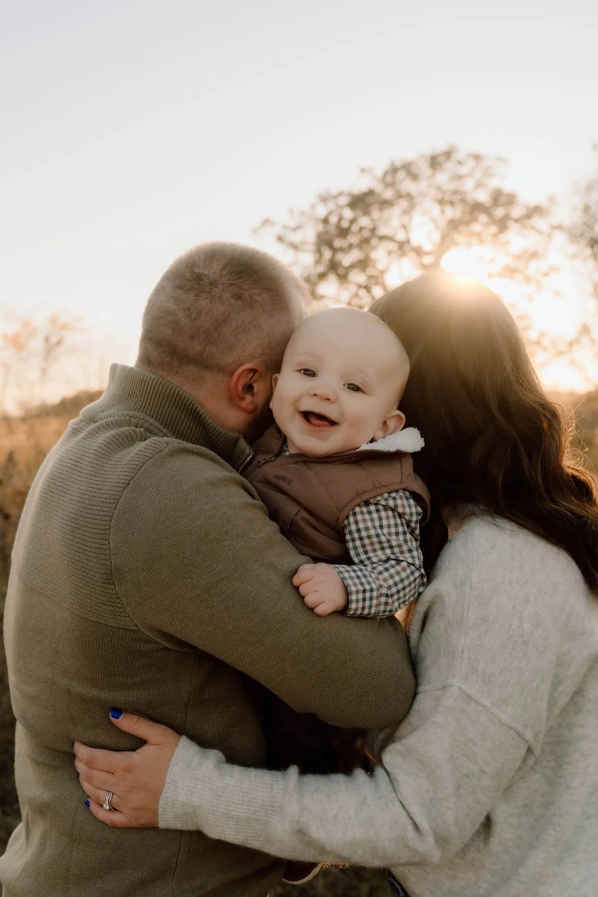 A happy family of three outdoors at sunset, with the father and mother holding their smiling baby boy between them.