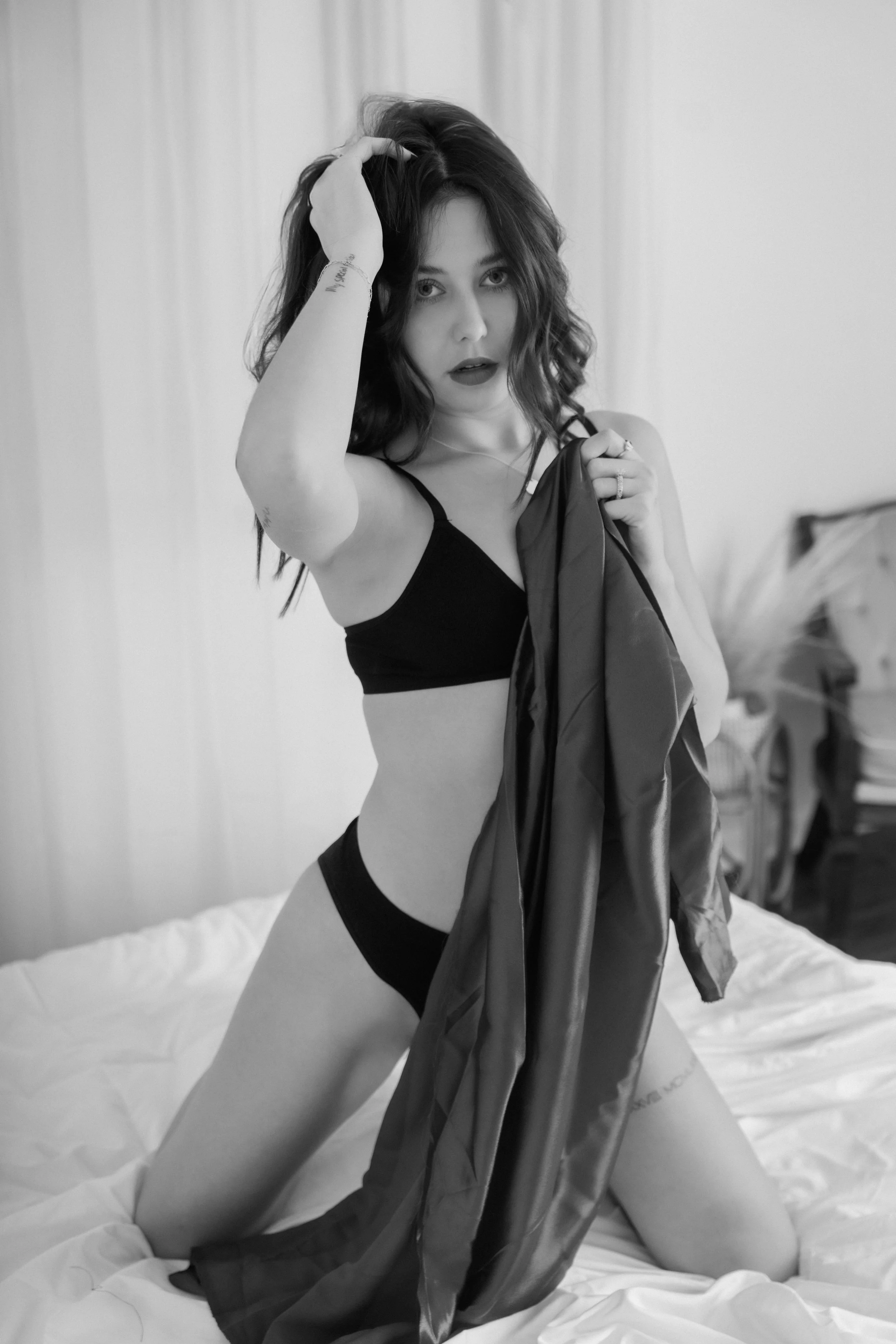 A woman with dark, wavy hair is kneeling on a bed, wearing black lingerie, holding a satin robe and running her fingers through her hair. The image is in black and white.