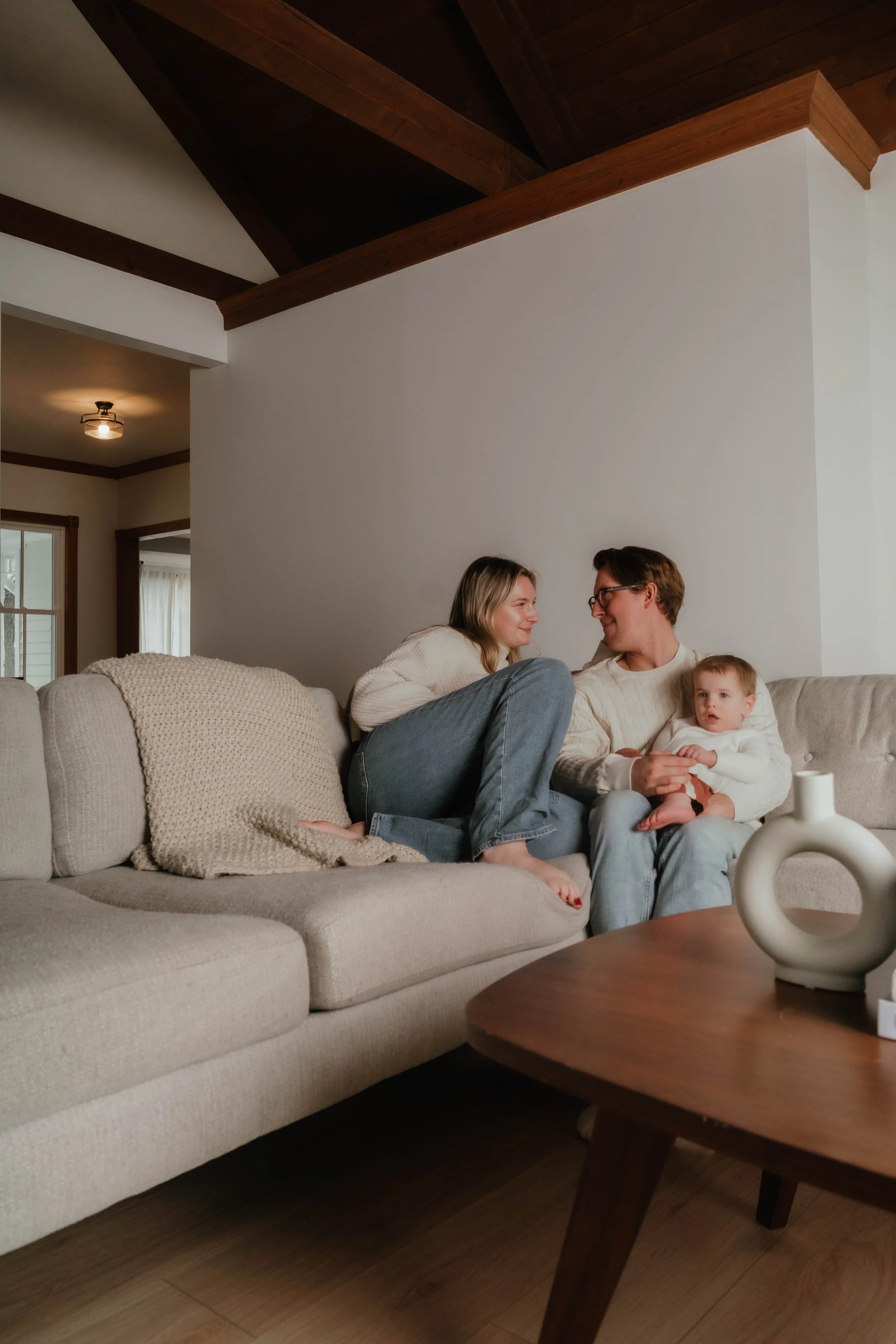 A couple and a young girl sitting together on a light-colored sofa in a cozy living room, with the girl holding a toy.