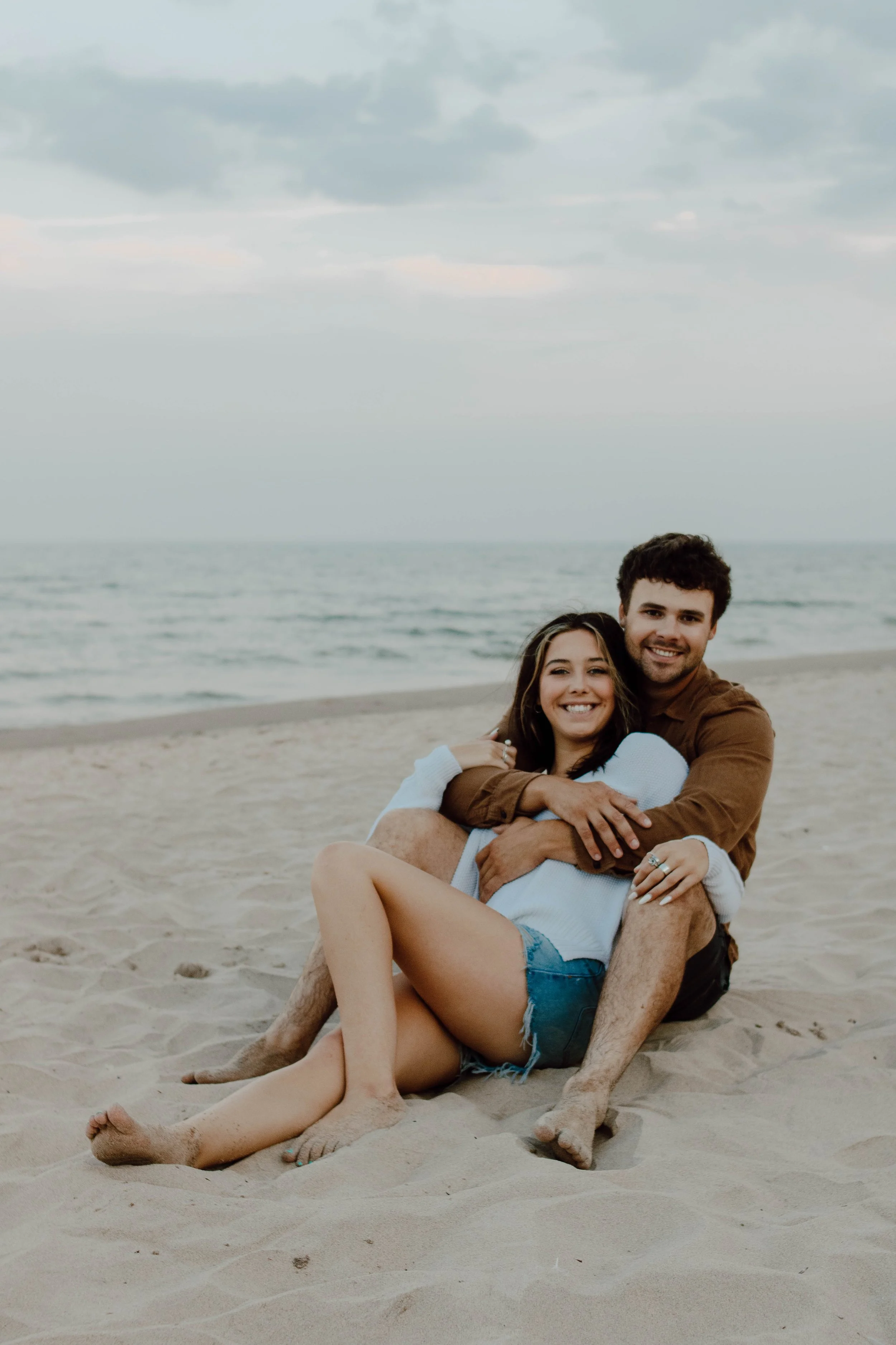 A young couple sitting on the sandy beach, embracing and smiling, with the ocean and cloudy sky in the background.