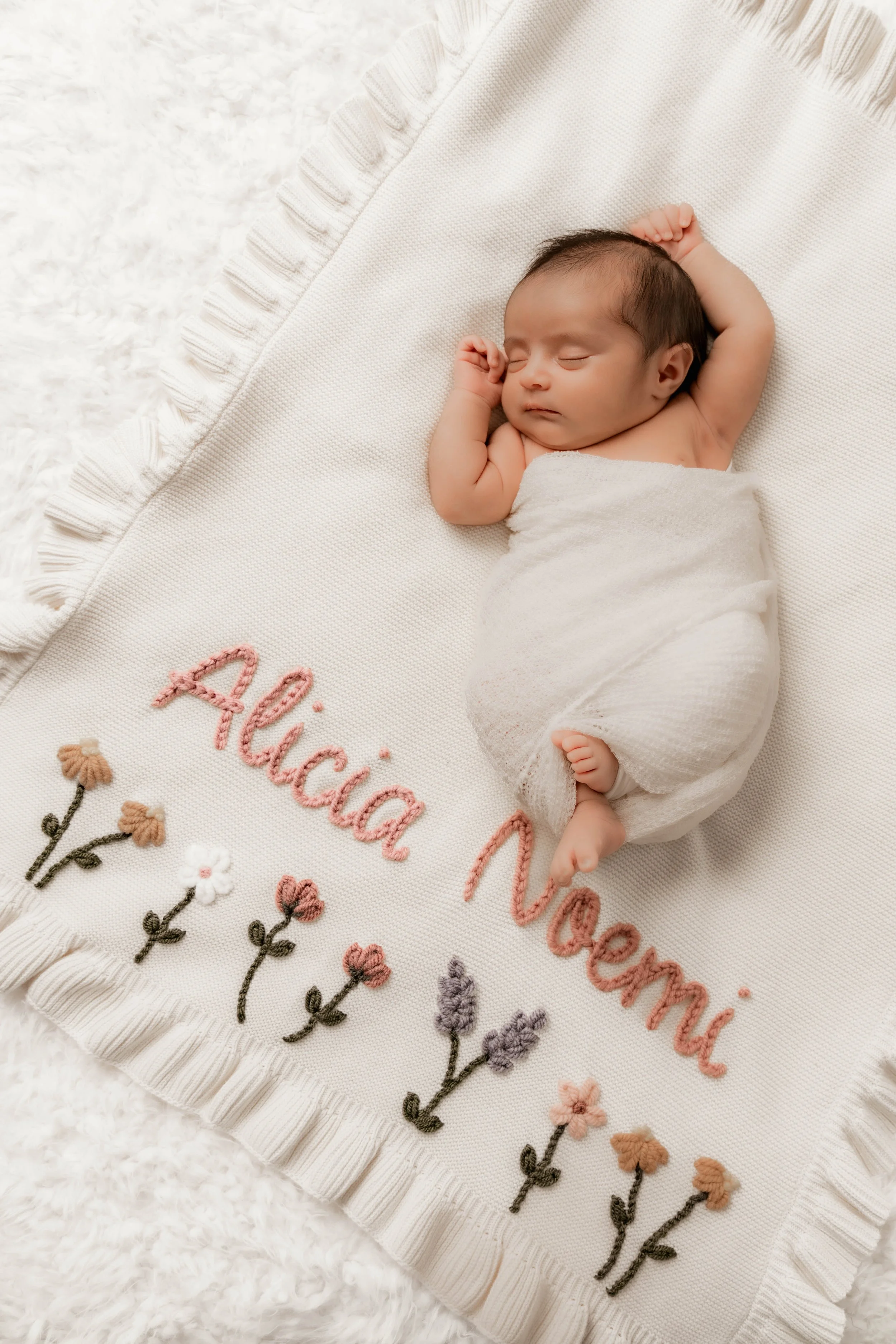 A sleeping baby wrapped in a white blanket, lying on a cream-colored blanket with embroidered pink and purple flowers and the name 'Alicia Noreen' in pink embroidery.