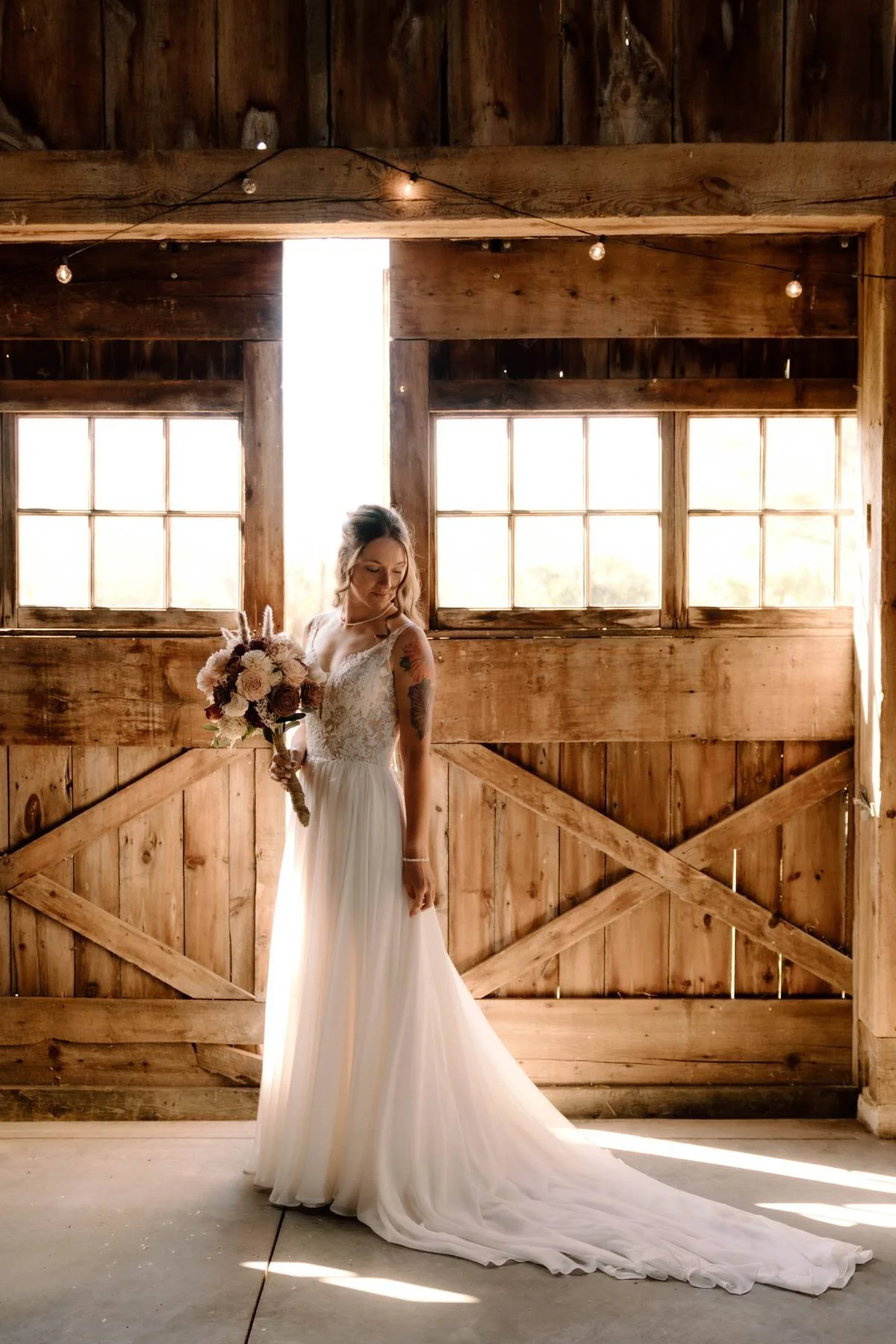 A bride in a white wedding dress holding a bouquet of flowers, standing in a rustic wooden room with sunlight streaming through windows behind her.