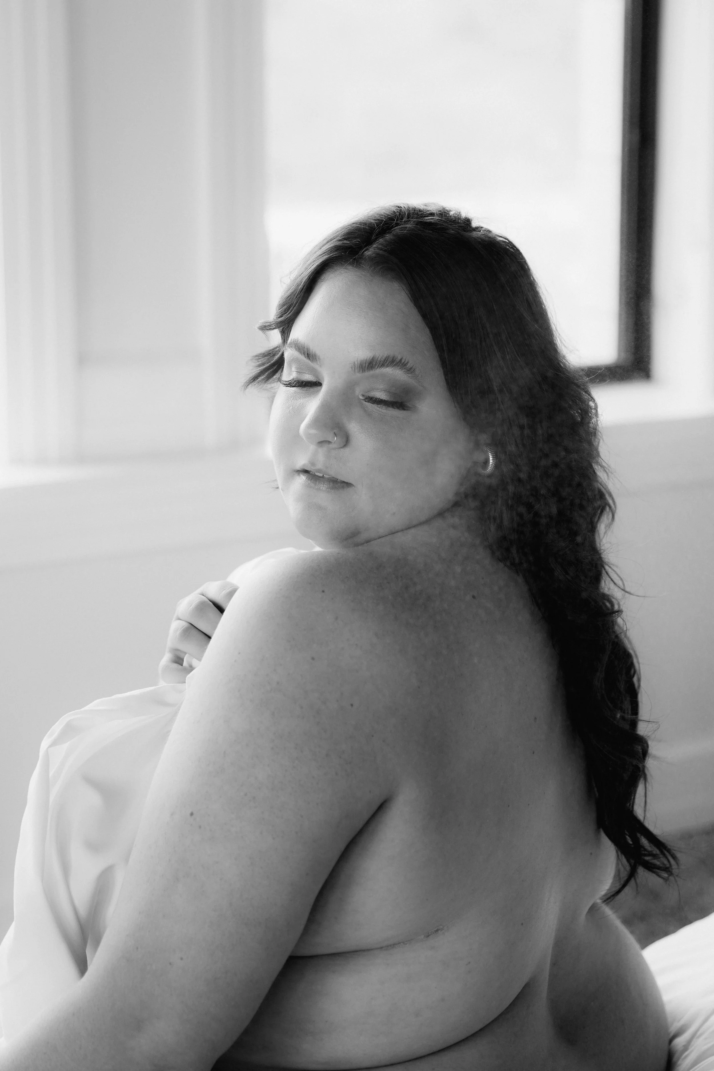 Black and white photo of a woman with closed eyes, shoulder-length hair, and a septum piercing, sitting topless with a sheet covering her chest, near a window with light filtering through.