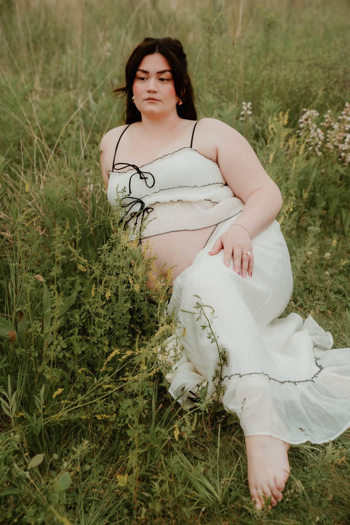 A woman with dark hair, wearing a white dress with black trim and spaghetti straps, is sitting in tall grass and wildflowers in a natural outdoor setting.
