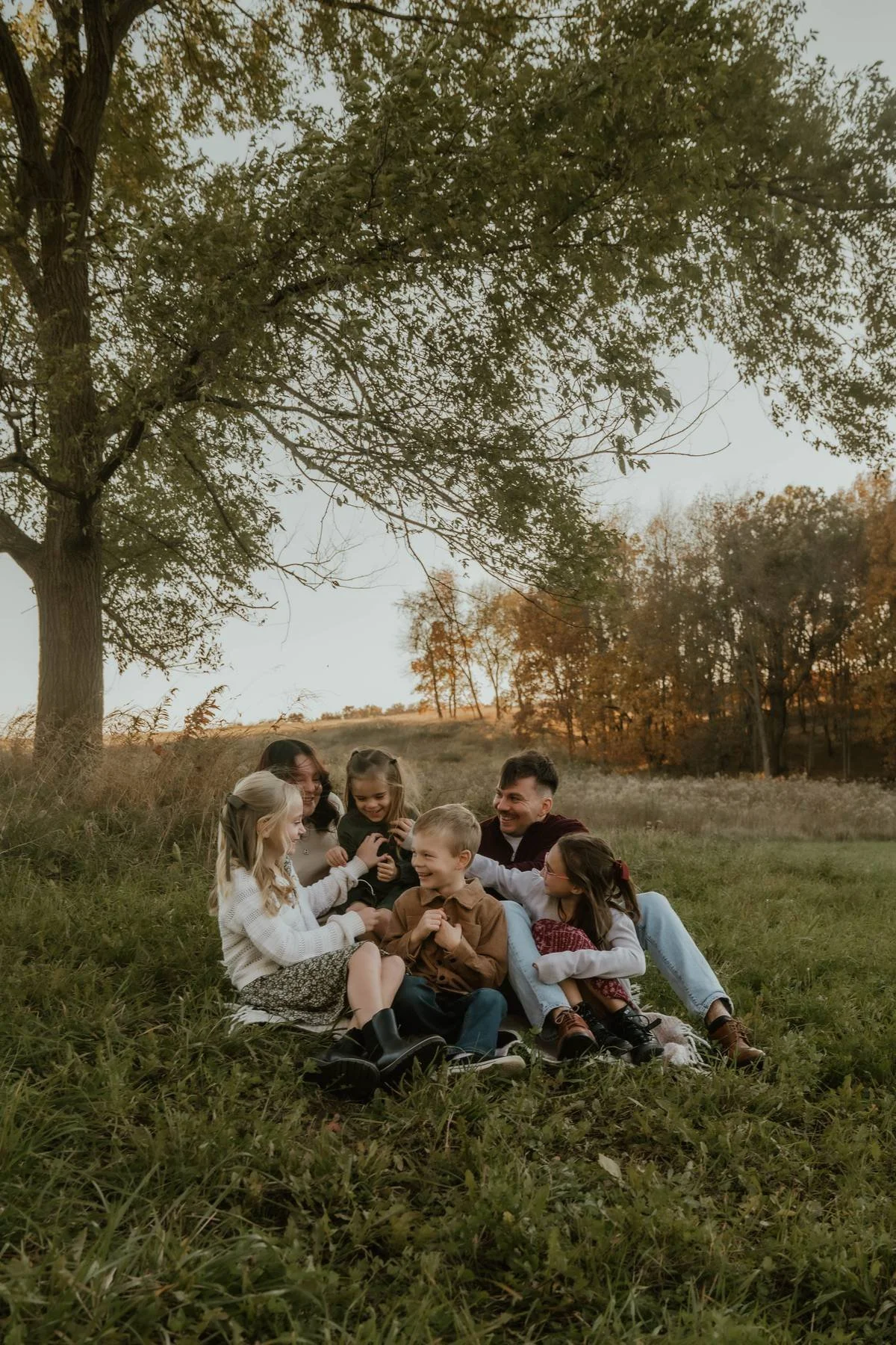 A family of six children and two adults sitting on the grass under a large tree in an open field, smiling and laughing together during sunset.