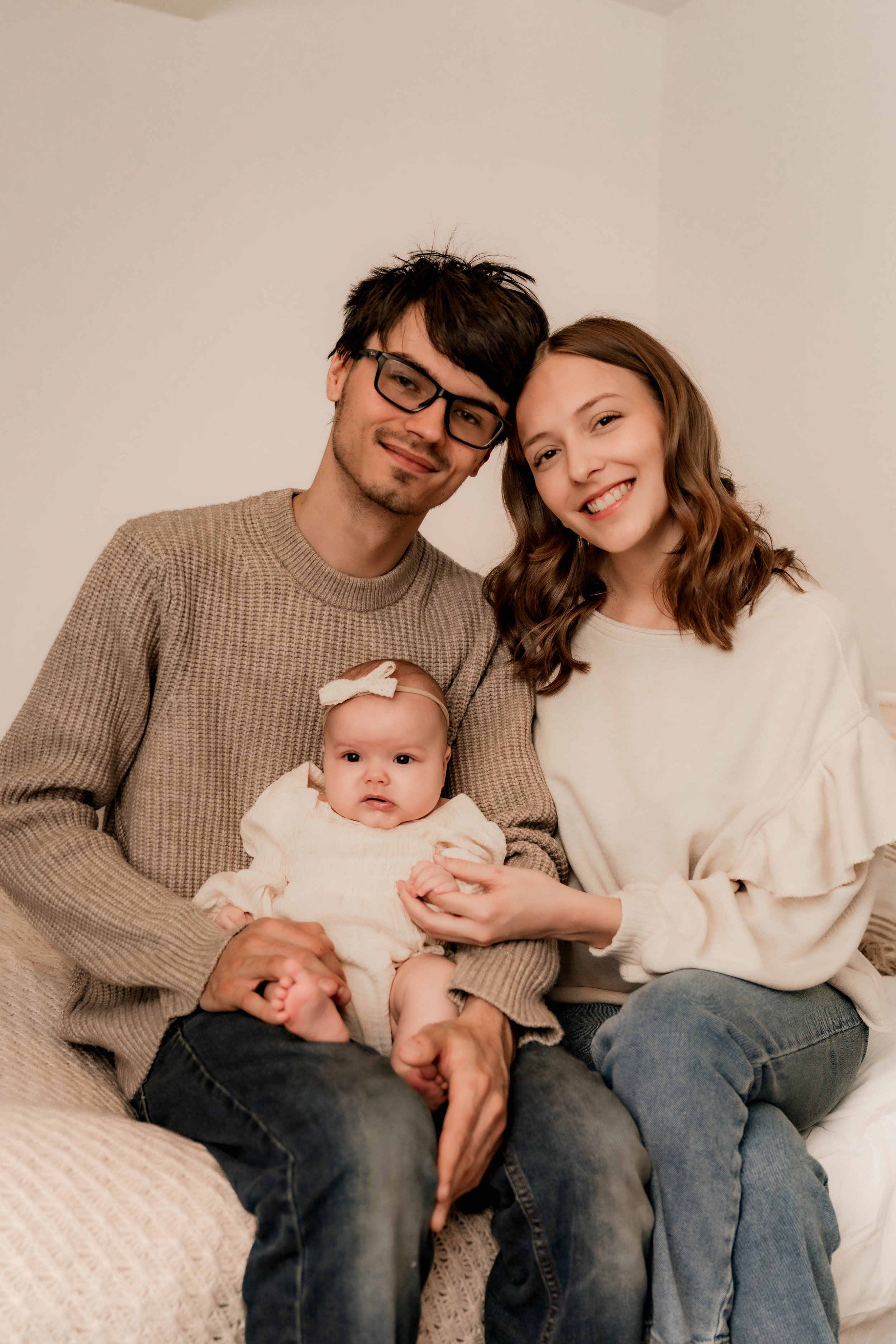 A family of three sitting together on a couch, with a man, woman, and baby girl. The man has dark hair, glasses, and is wearing a beige sweater, holding the baby. The woman has wavy brown hair, a light-colored sweater, and is smiling. The baby girl i
