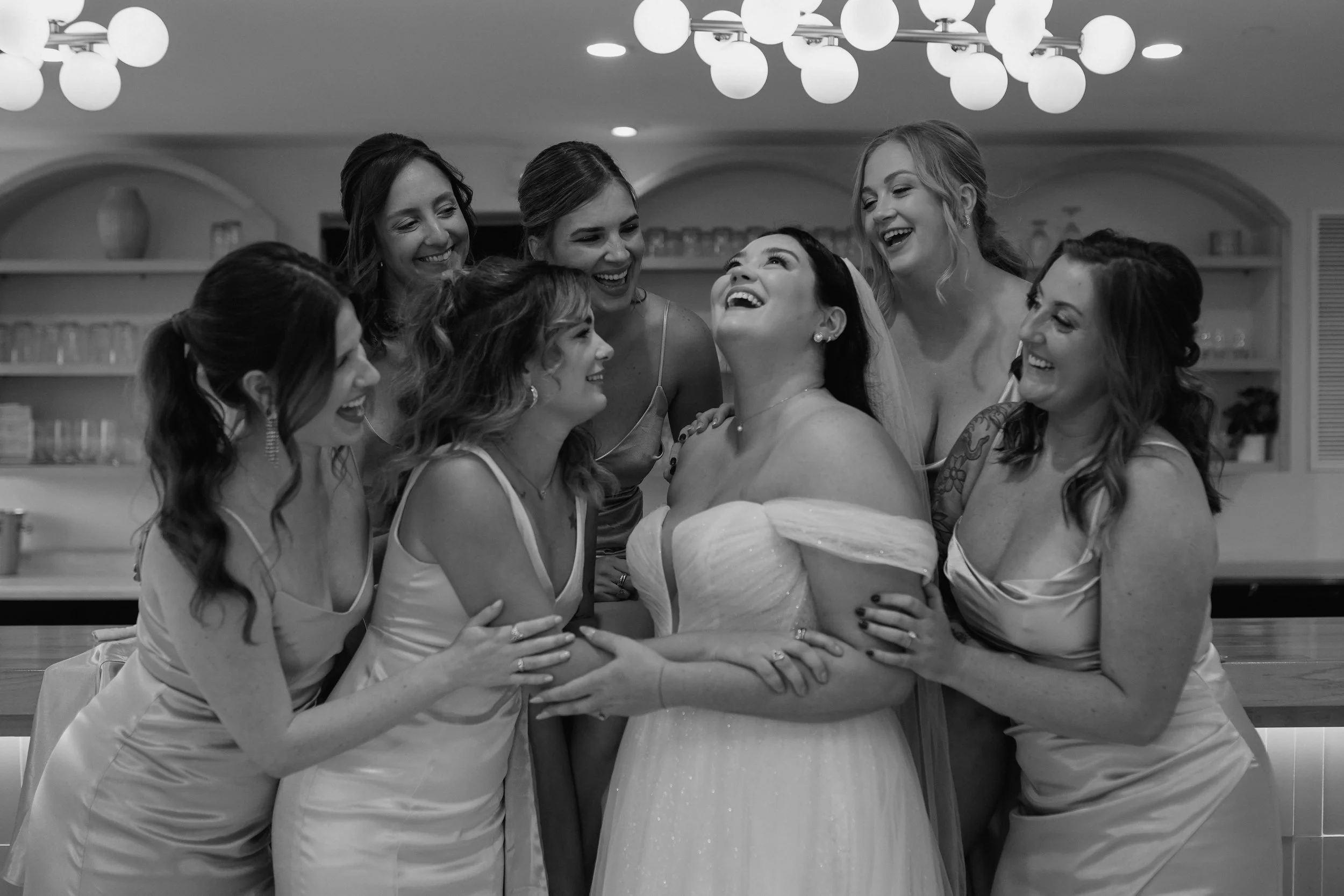A bride and her bridesmaids laughing and hugging each other at a wedding celebration indoors.