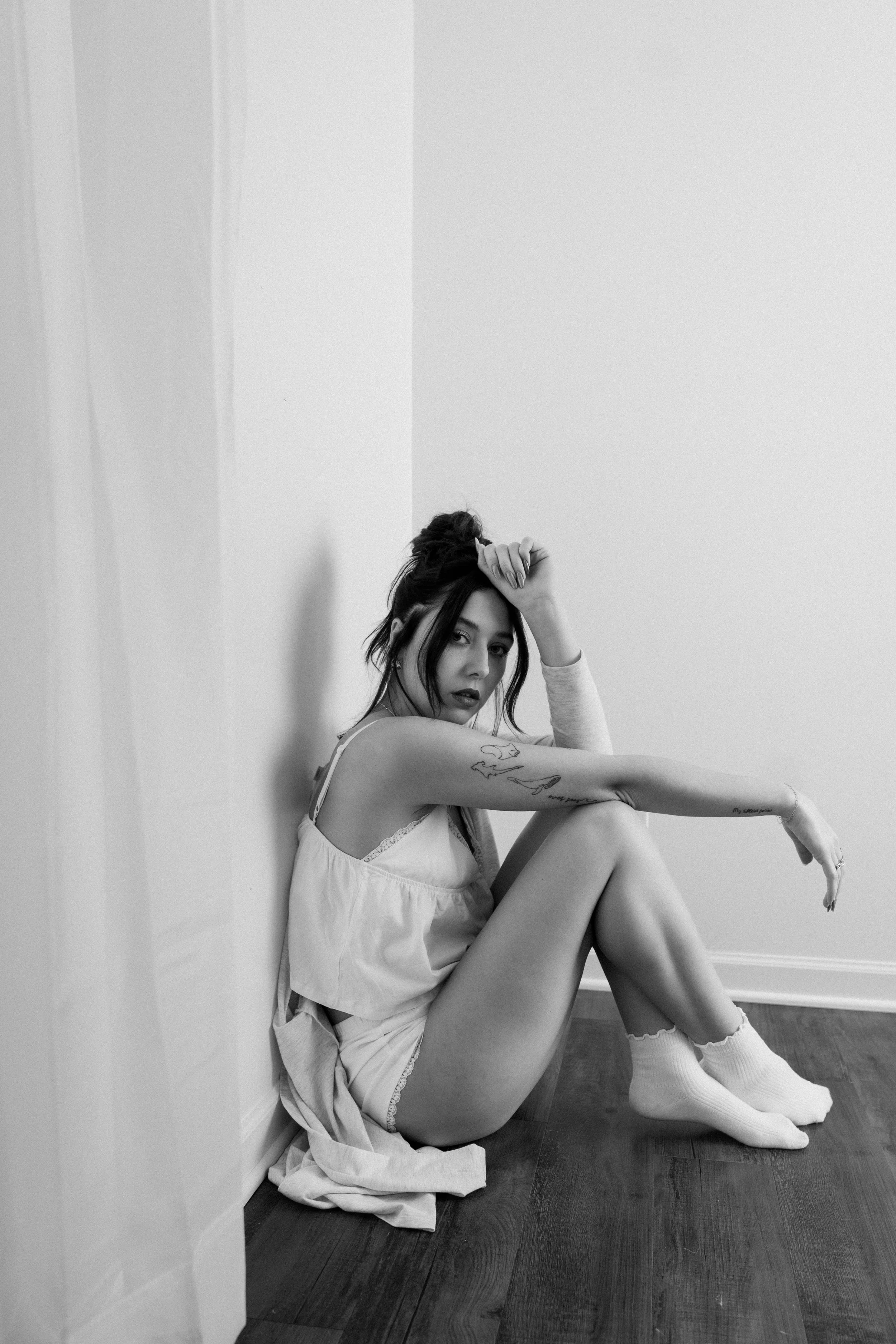 A young woman with dark hair in a messy bun, sitting on the wooden floor in a corner of a room. She is wearing a light-colored nightgown, socks, and has tattoos on her arm. She has a contemplative expression and is resting her head on her hand, with 