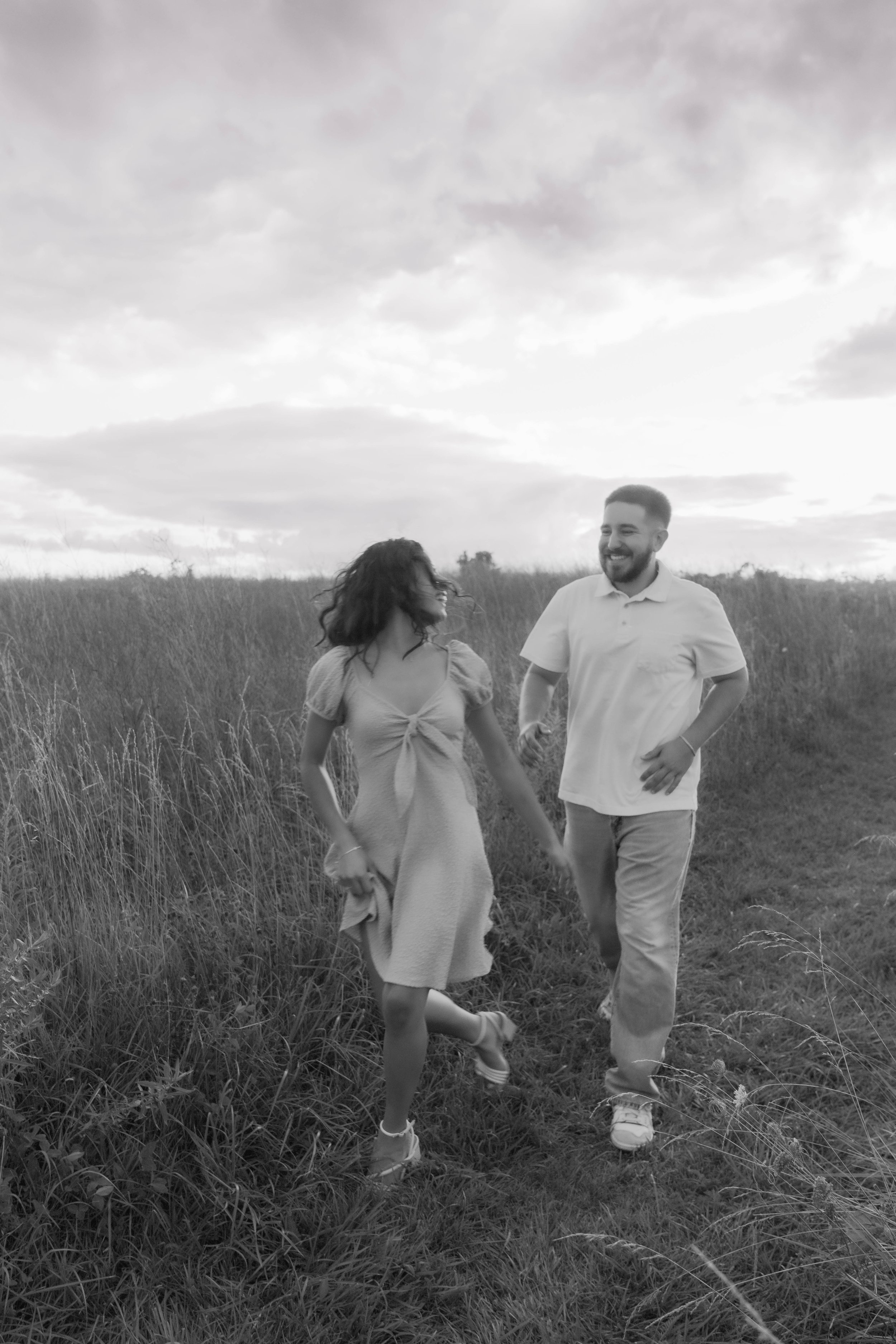 A black and white photo of a smiling man and woman walking hand in hand through a grassy field during sunset or sunrise.