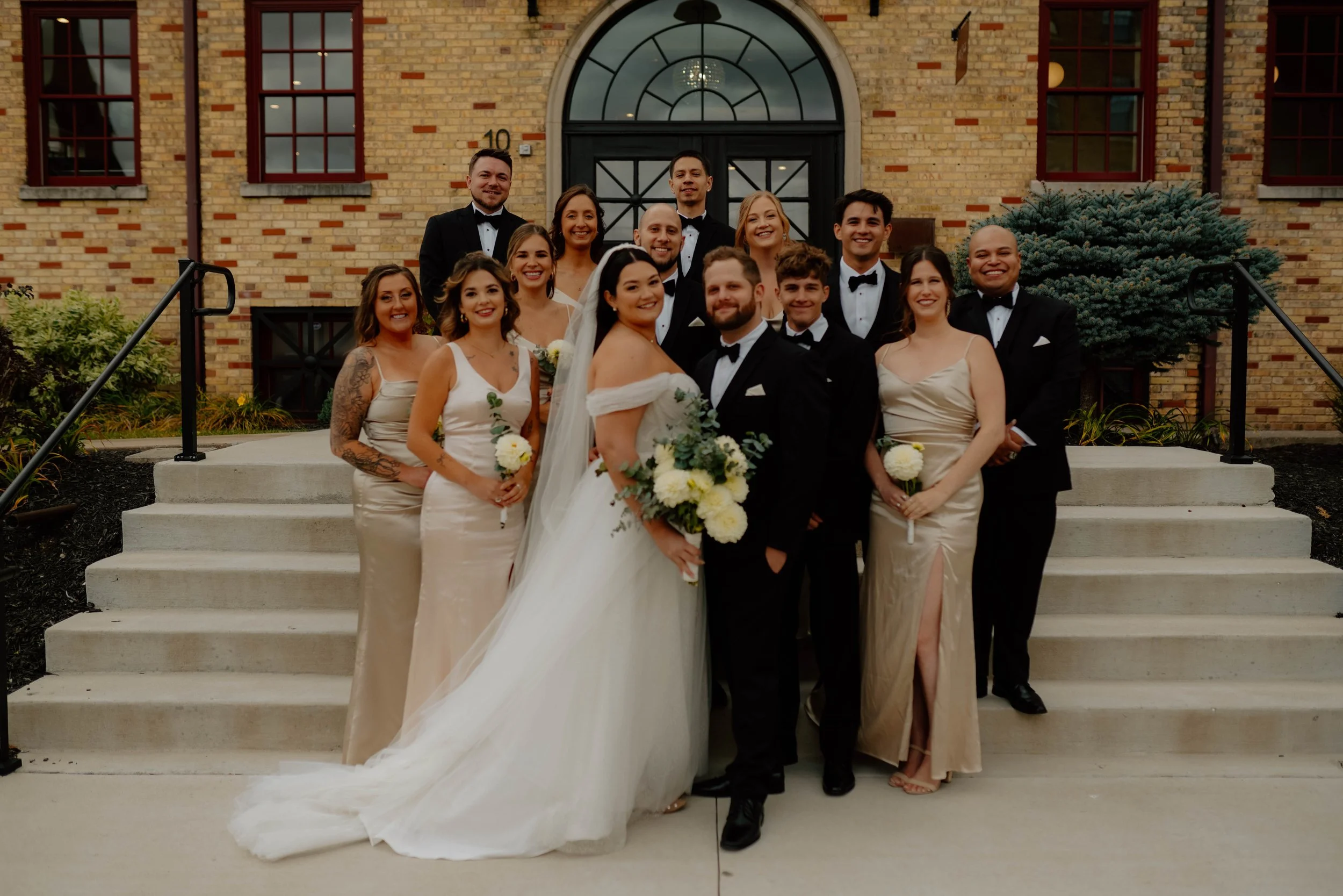 Group photo of wedding party with bride, groom, bridesmaids, and groomsmen standing on steps in front of a brick building with large arched window and greenery.