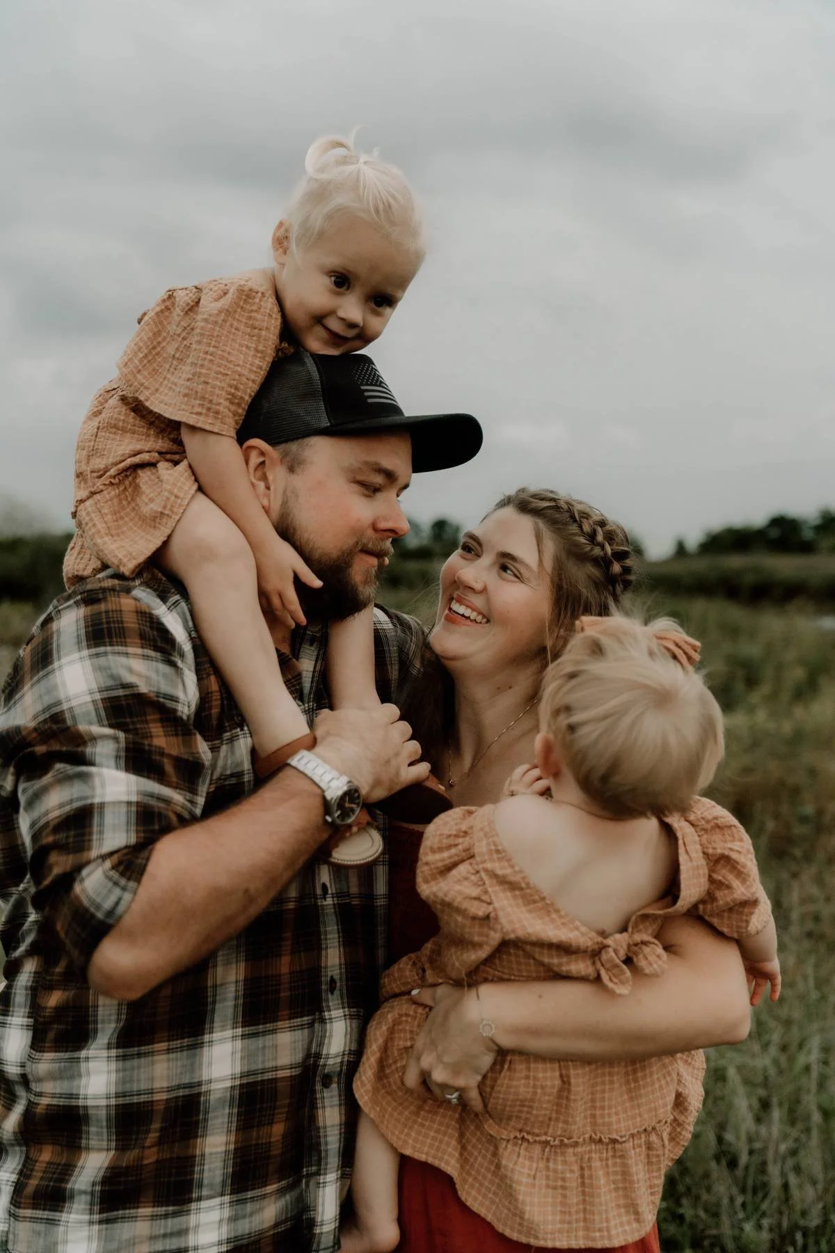 A family of four outdoors, with two children being held by their parents, enjoying a moment on a cloudy day.