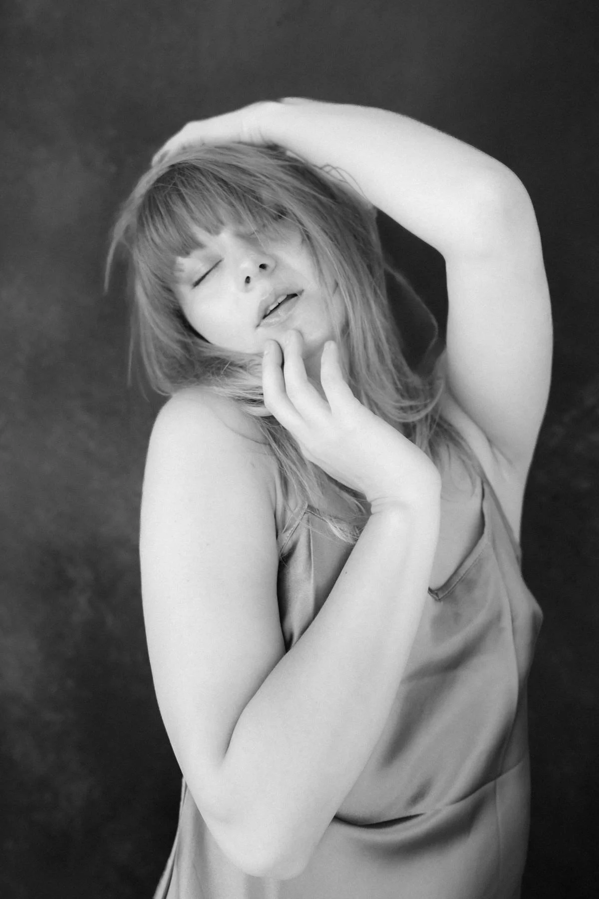 Black and white photograph of a woman with closed eyes, tousled hair, and her hand touching her face, posing with one arm raised above her head.