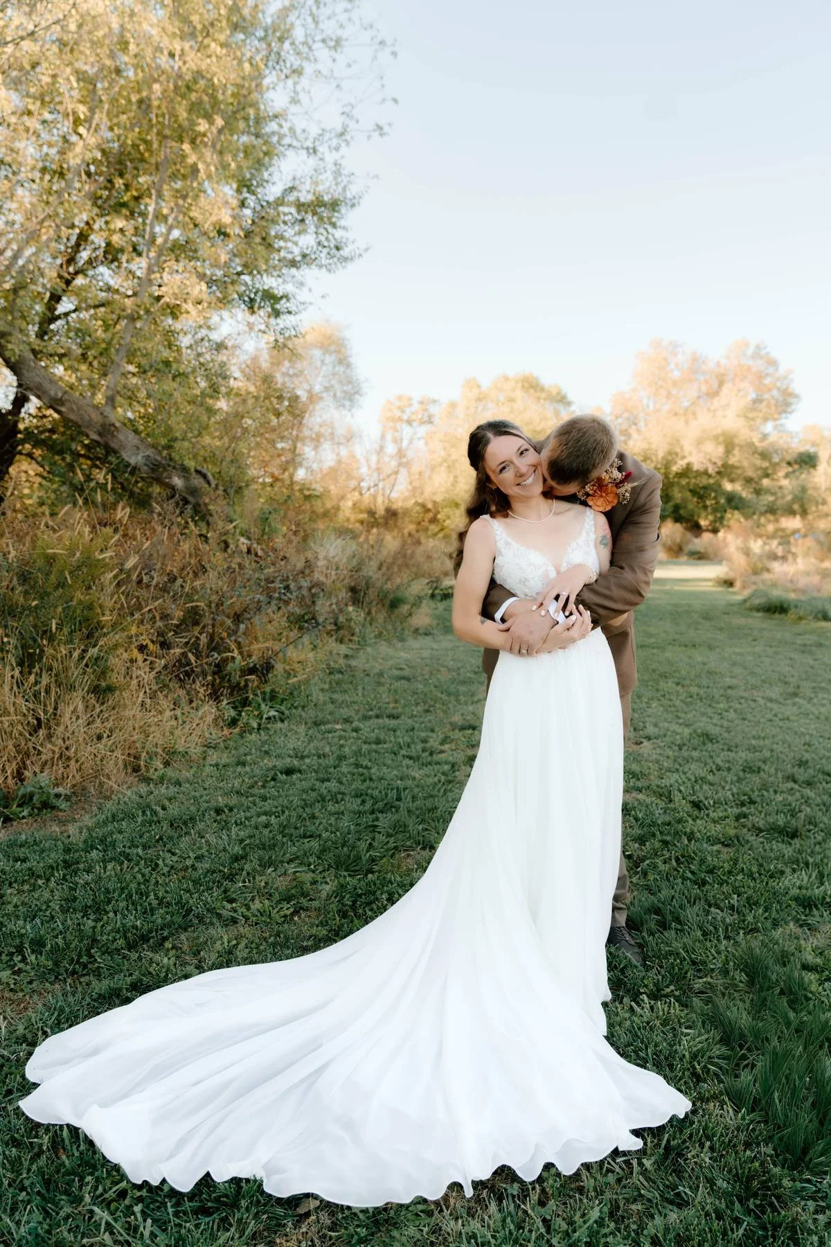 A happy bride in a white wedding dress with a train, smiling while a man in a brown suit kisses her cheek in an outdoor setting with trees and grass.