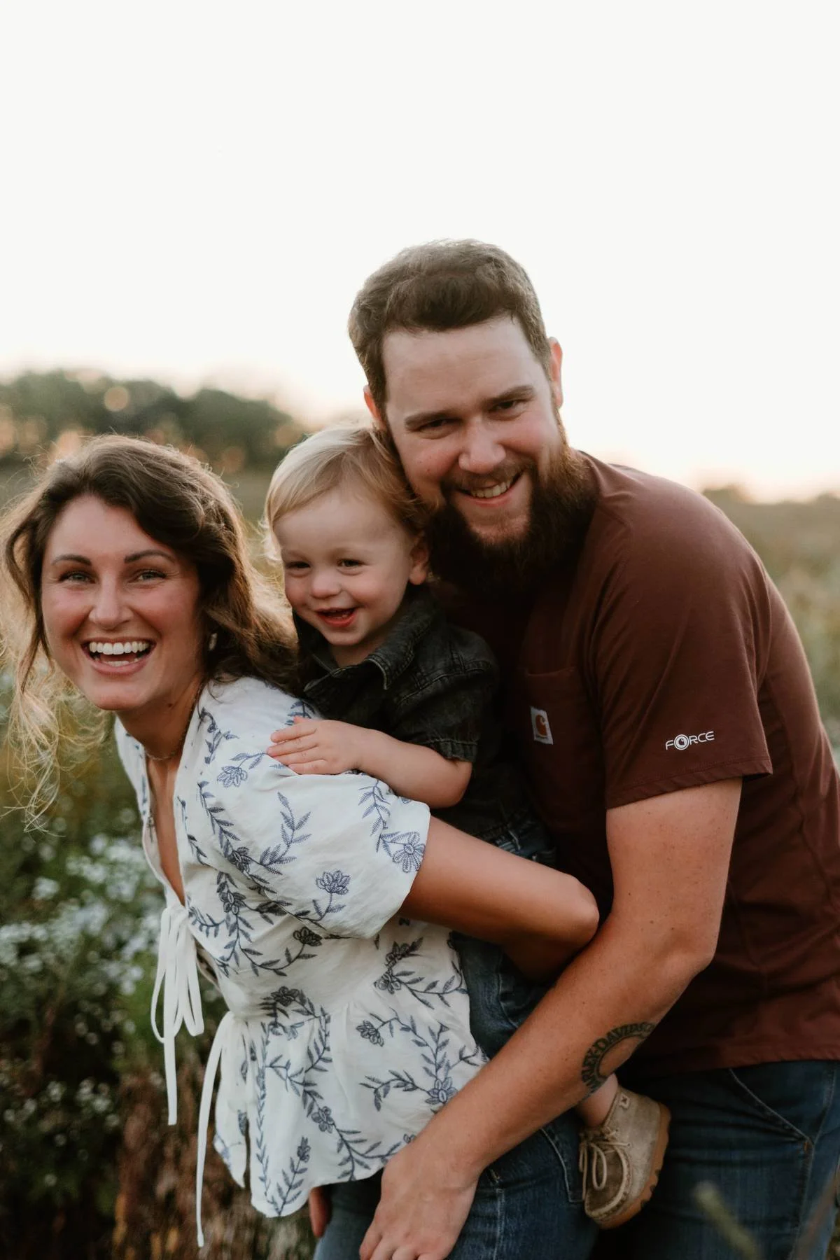A happy family of three outdoors in a field during sunset, smiling and embracing each other.