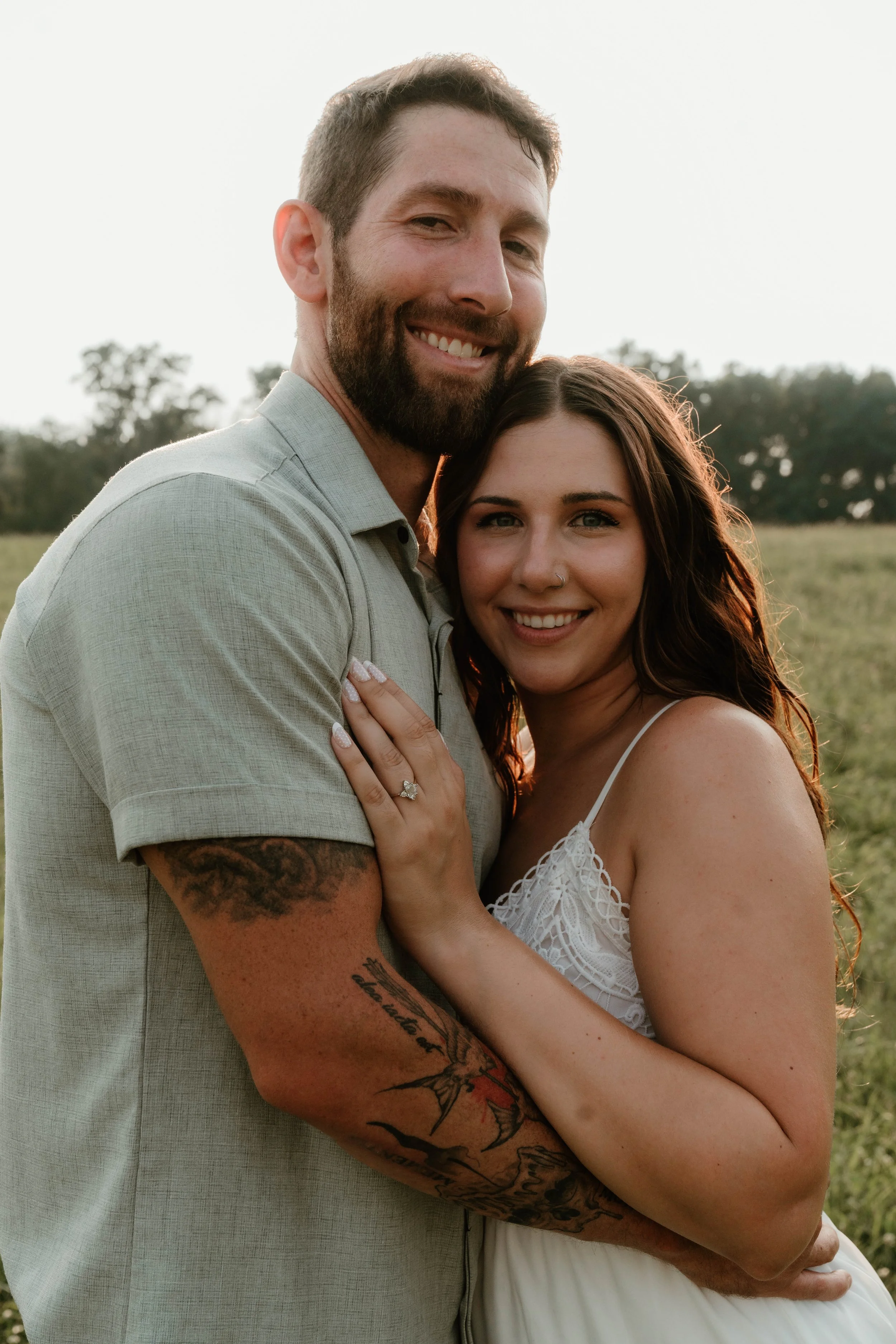 A smiling couple embracing outdoors in a field during sunset, woman wearing a white spaghetti strap dress with a ring on her finger, man with a beard, tattoos on his arm, wearing a light-colored shirt.