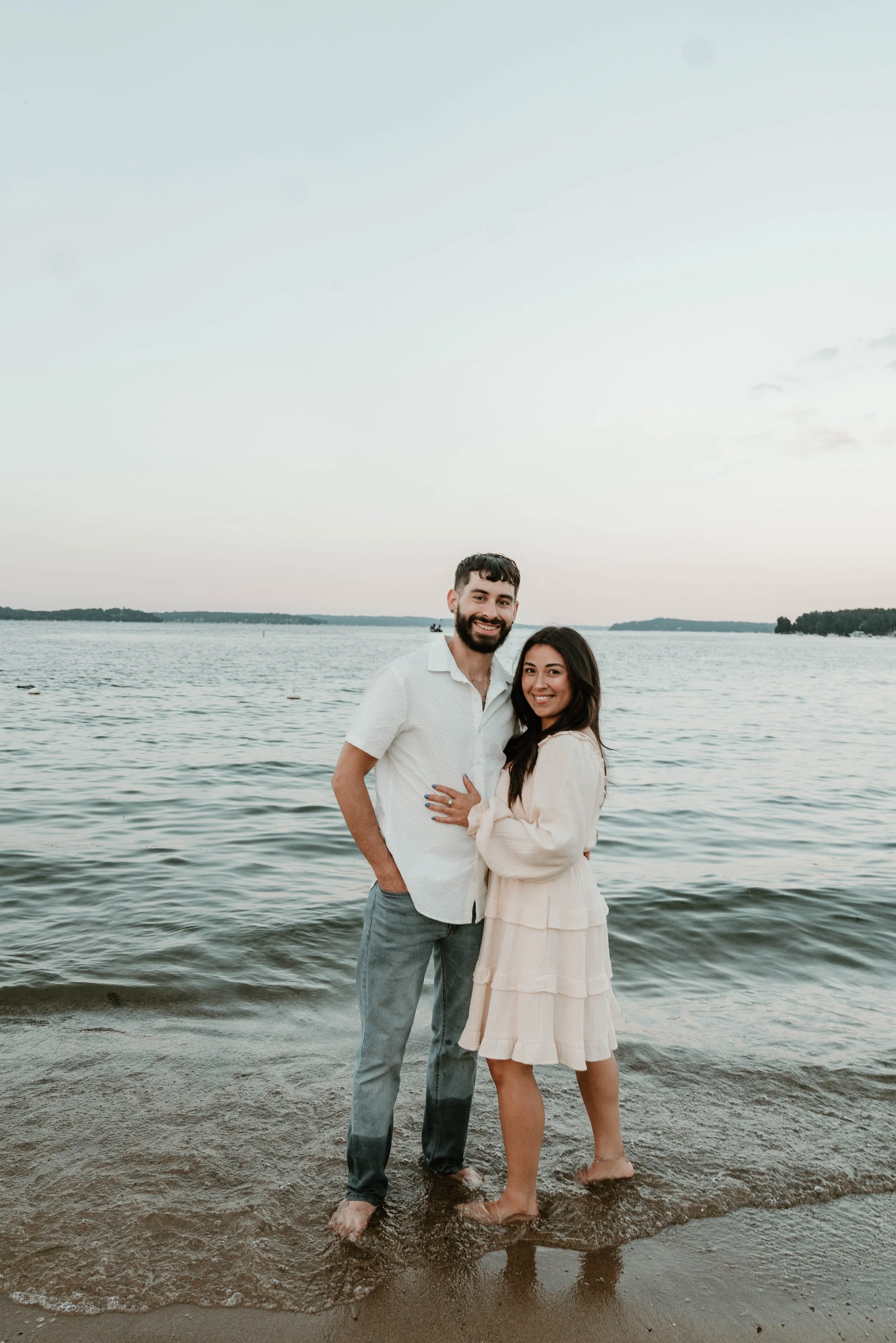 A happy couple stands barefoot on a beach near the water, smiling at the camera during sunset.