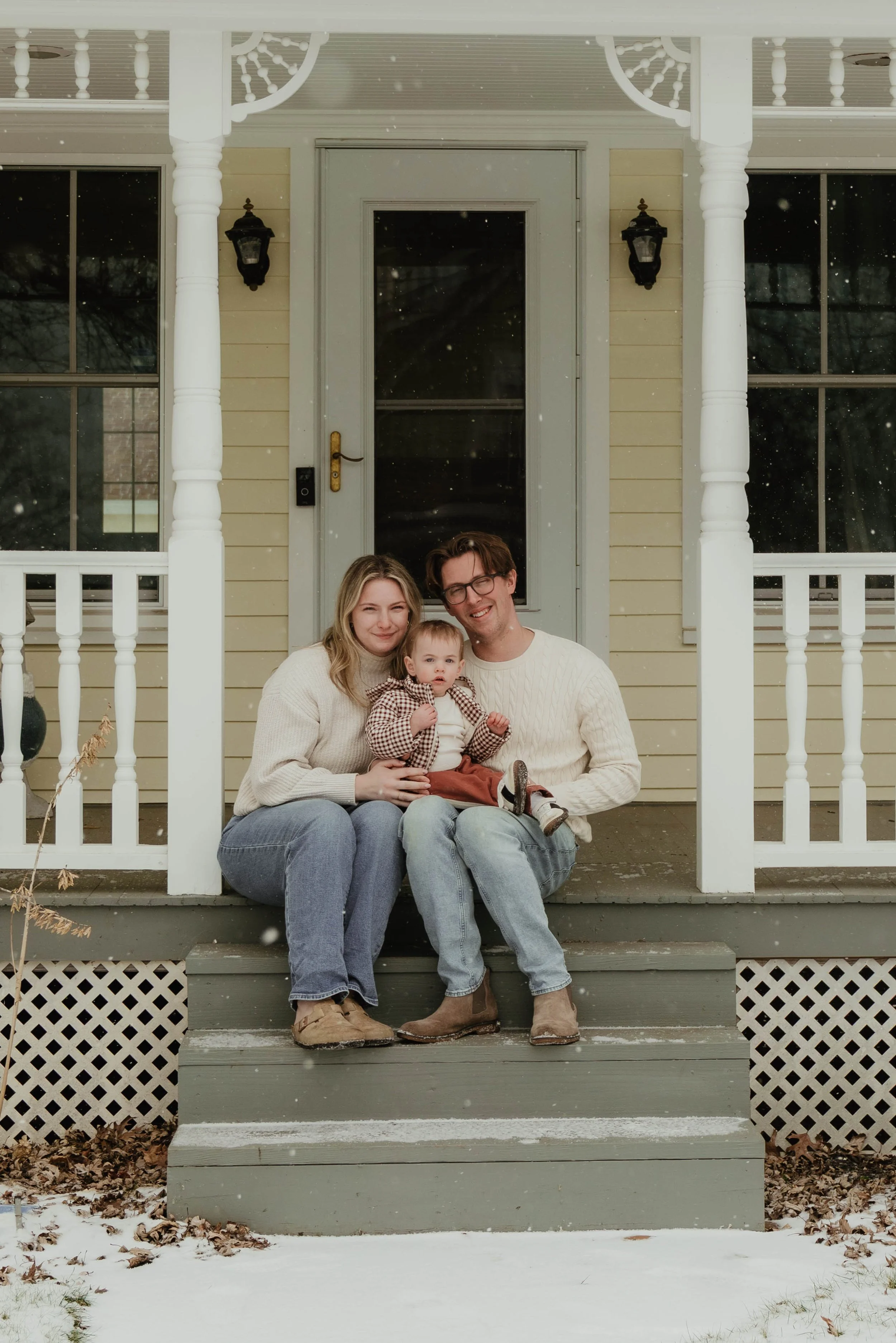 A family of three sitting on the front porch of a house during winter, with snow on the ground and falling snowflakes. The family includes a woman, a man, and a young child, all smiling and dressed warmly.
