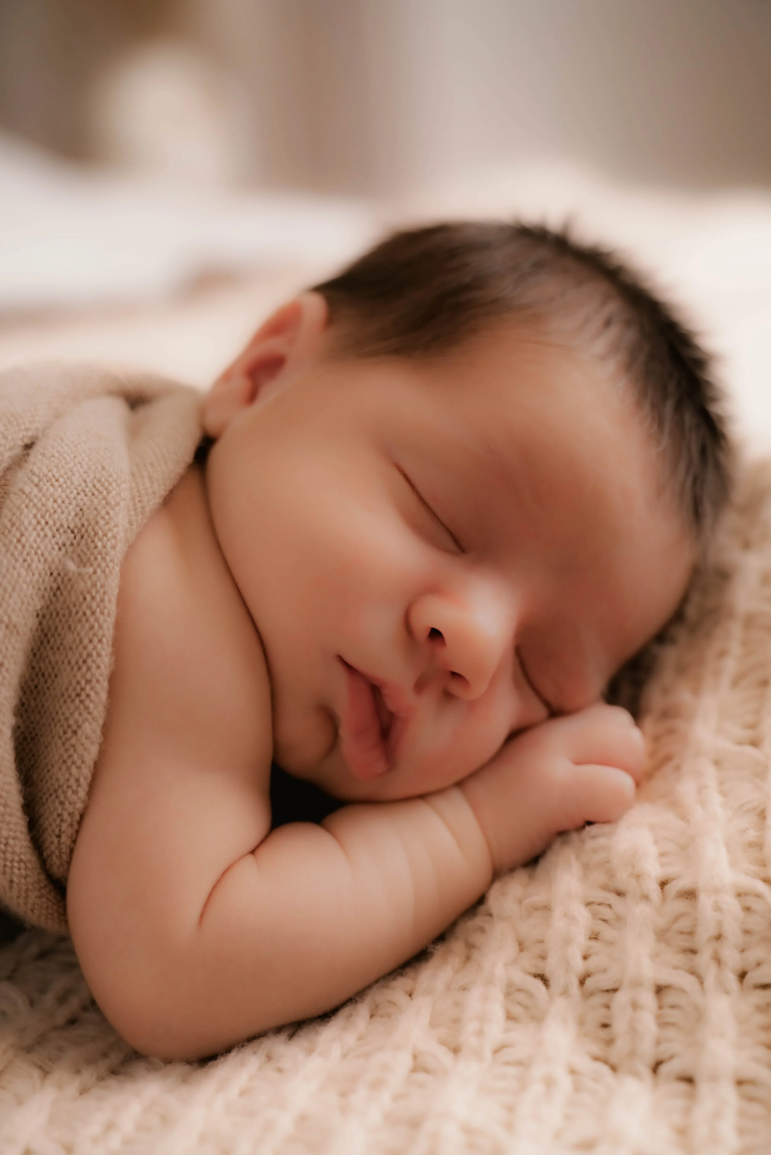Close-up of a sleeping baby lying on a knitted blanket, with eyes closed and hand resting near the face.