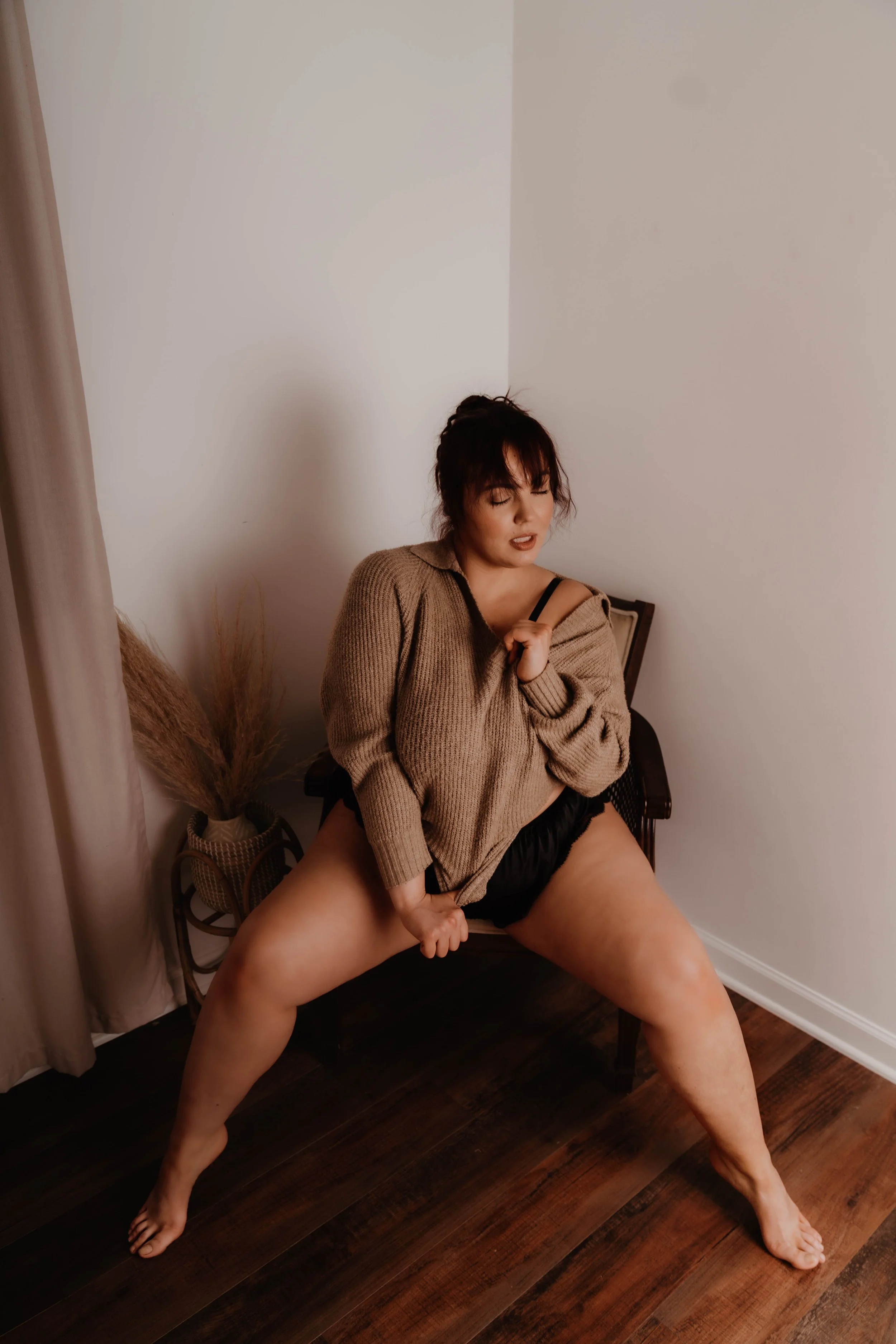 A woman crouches on a chair in a corner of a room, wearing a beige sweater and black shorts, with her legs spread apart and one hand gripping her shorts. She has dark hair with bangs and appears to be posing confidently.