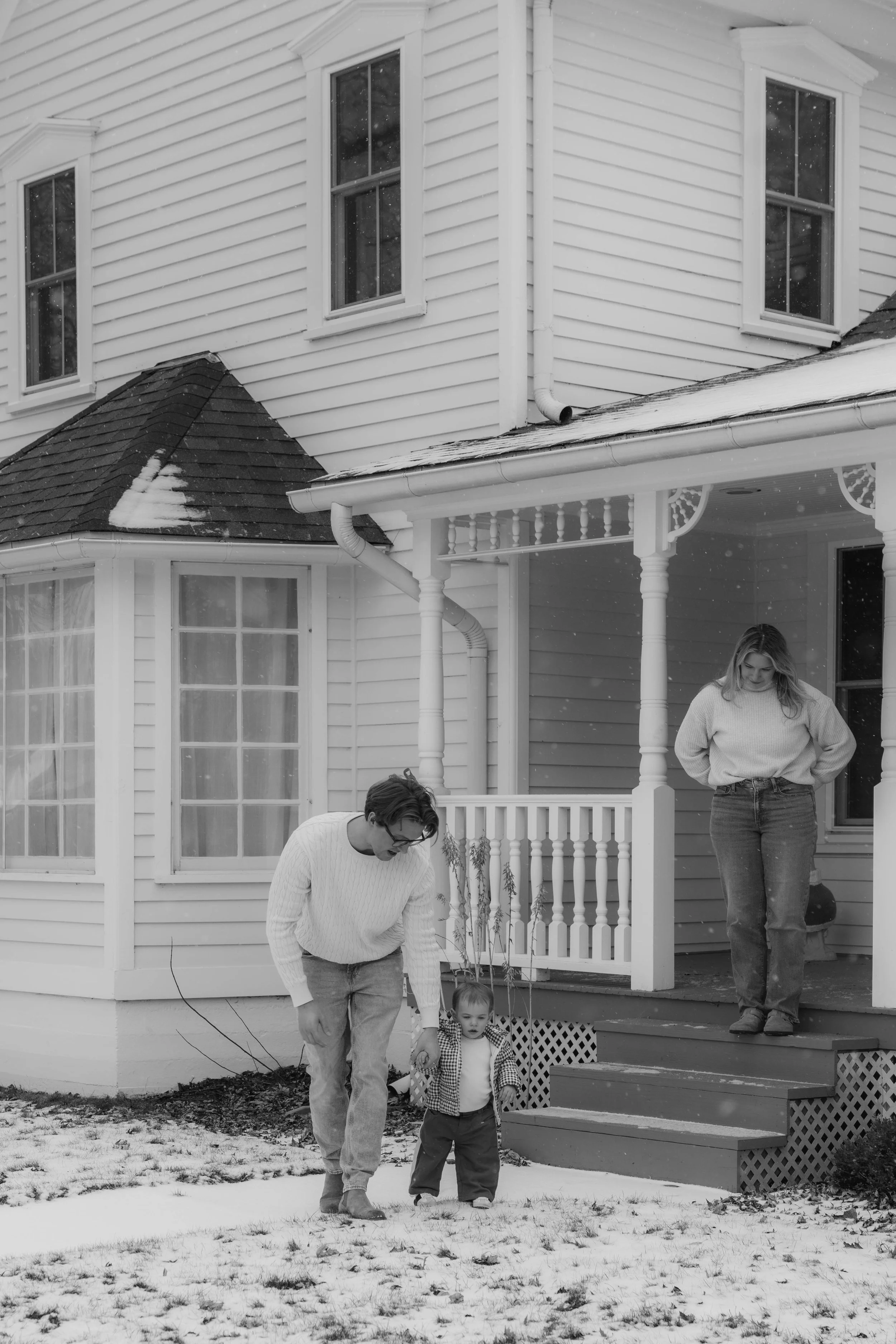 A family standing outside a white house in winter, with snow on the ground and on the roof. A young man and woman are with a small child on the front steps, the man holding the child's hand as they walk on the snow.
