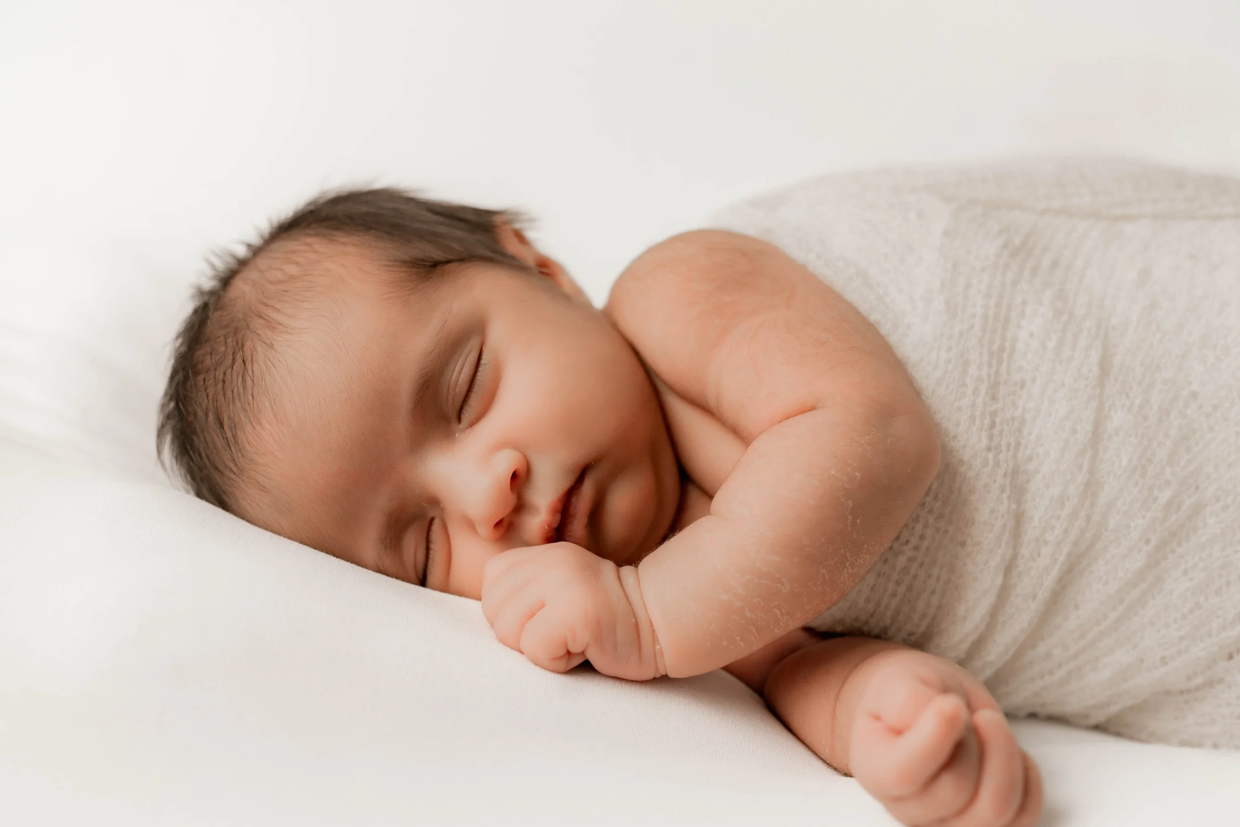 A sleeping baby with dark hair, lying on a white surface, wrapped in a light-colored blanket.