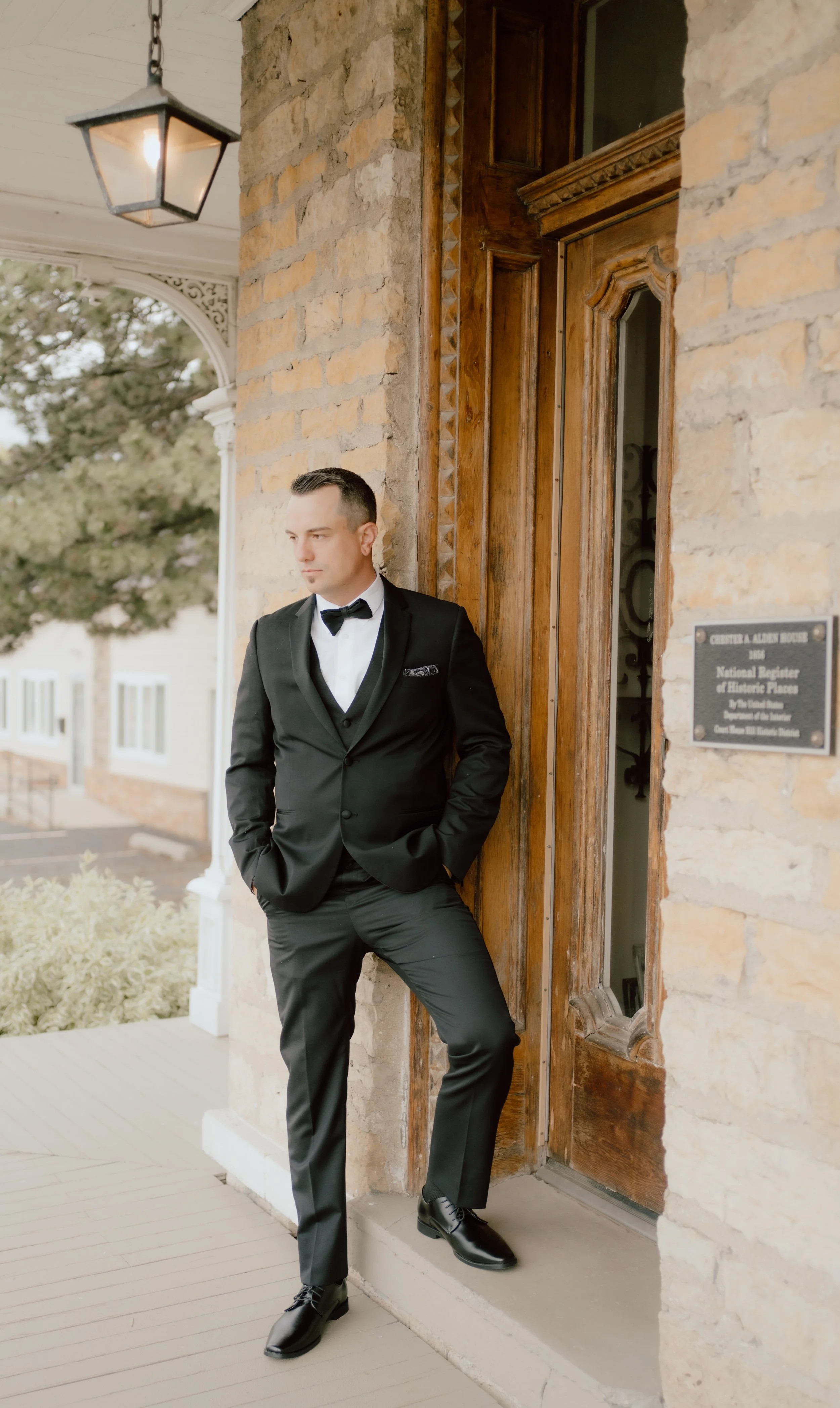 A man in a black tuxedo leaning against a wooden doorframe on a building with a brick exterior.