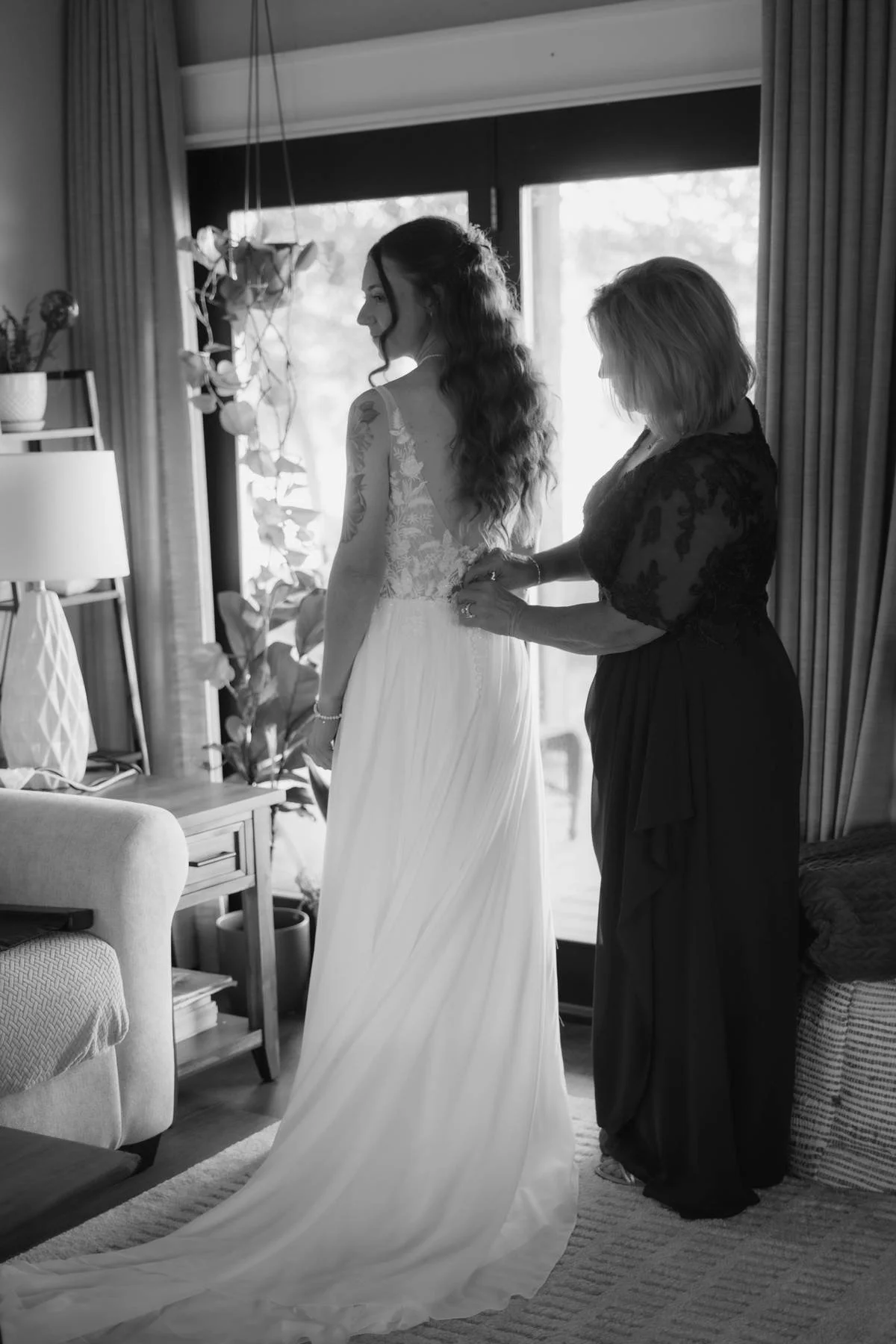 A bride in a wedding dress is assisted by a woman, possibly her mother, as she prepares for her wedding in a cozy living room with large windows, curtains, and indoor plants.
