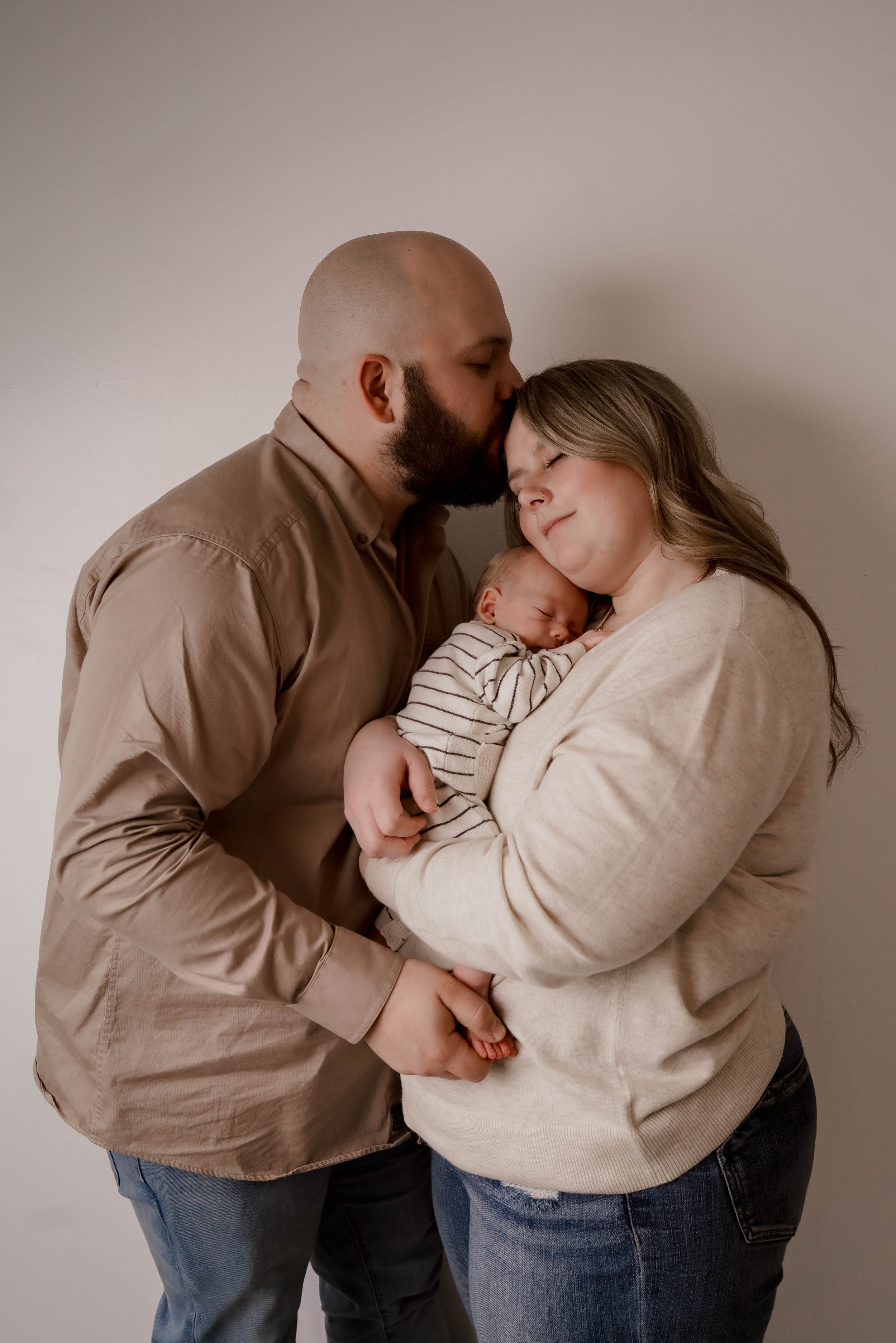 A family of three: a man, woman, and baby, standing close together, with the man gently kissing the woman's forehead as she holds the newborn, all smiling softly.