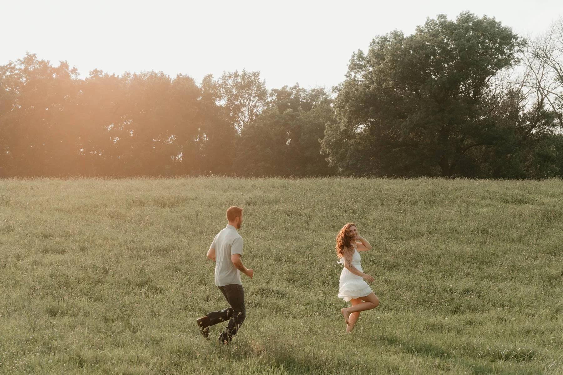 A man and woman running and playing in a grassy field during sunset, with trees in the background.