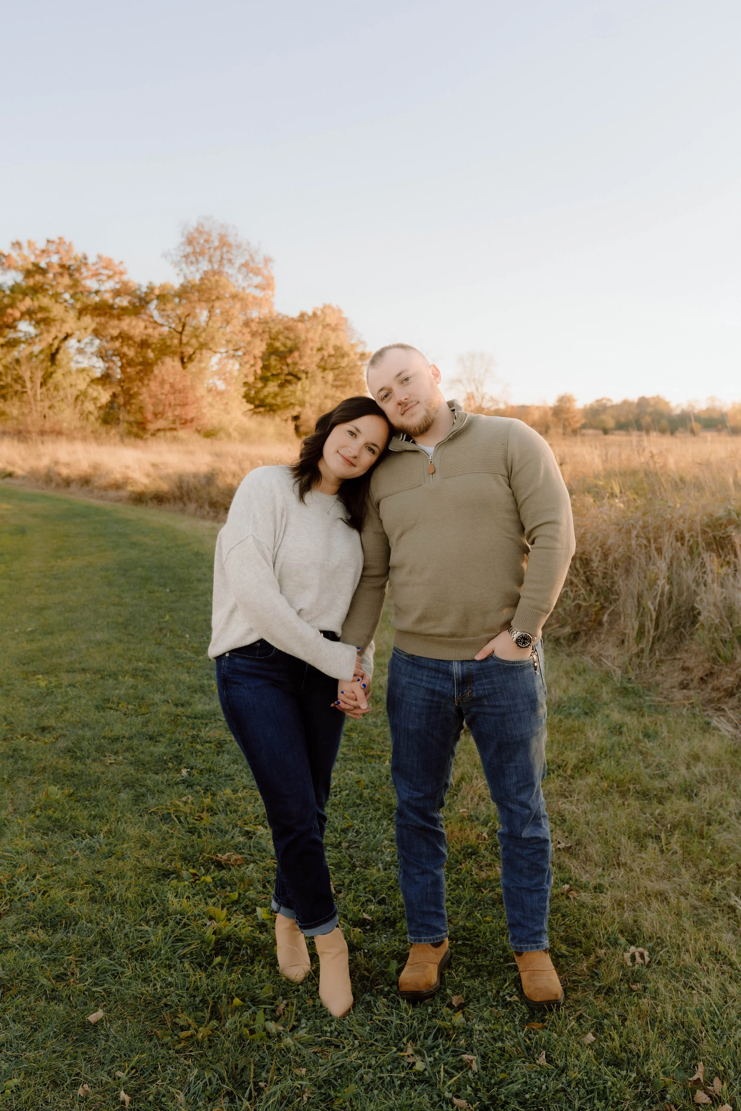A young couple standing outdoors in a grassy field during autumn, holding hands and leaning their heads together, with trees showing fall foliage in the background.