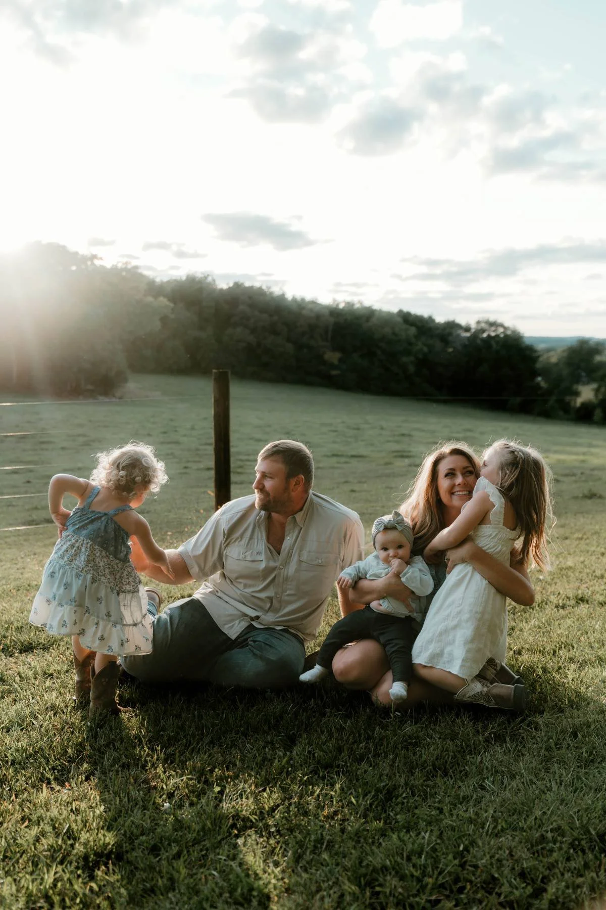 A happy family of five outdoor during sunset on the grass, sitting and embracing. The father, mother, and three young daughters are smiling and interacting, with trees and cloudy sky in the background.