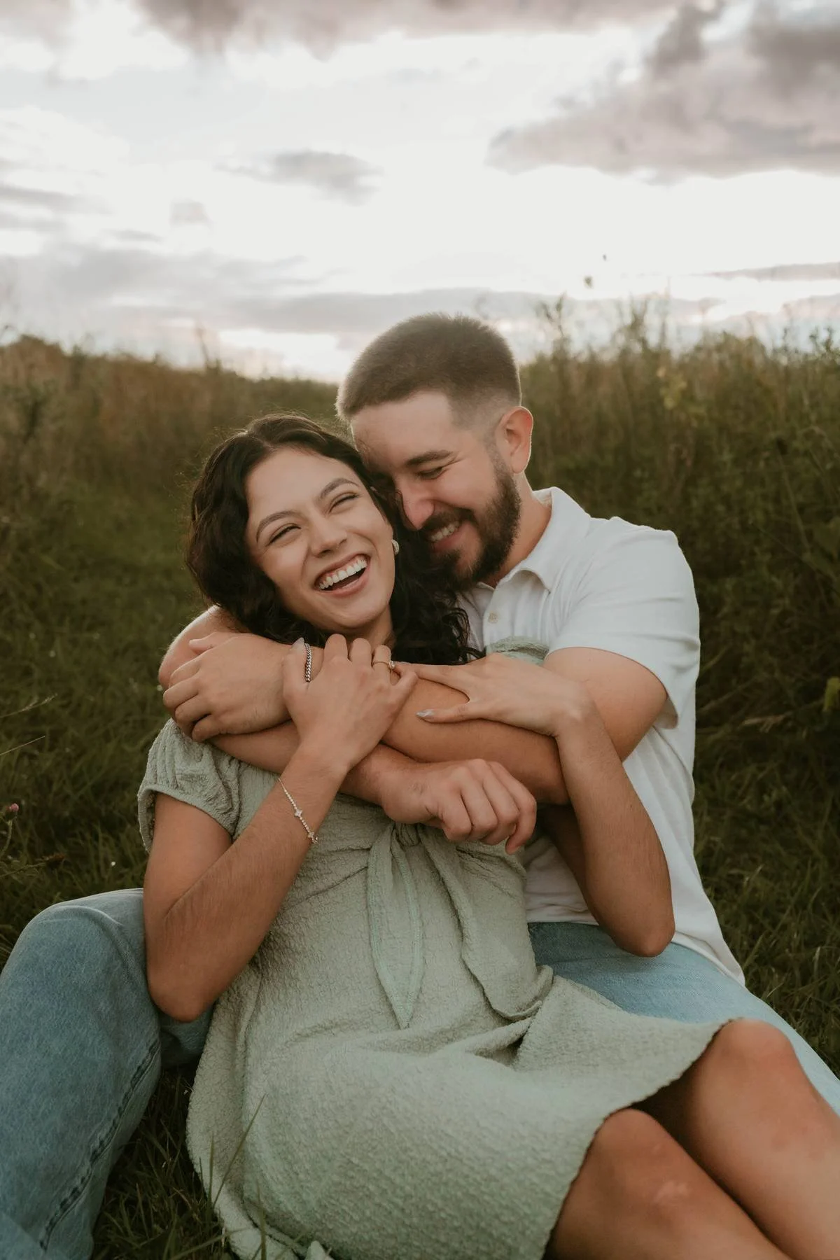 A happy couple sitting outdoors embracing, with the woman smiling broadly and the man nuzzling her cheek, during sunset on a grassy field.