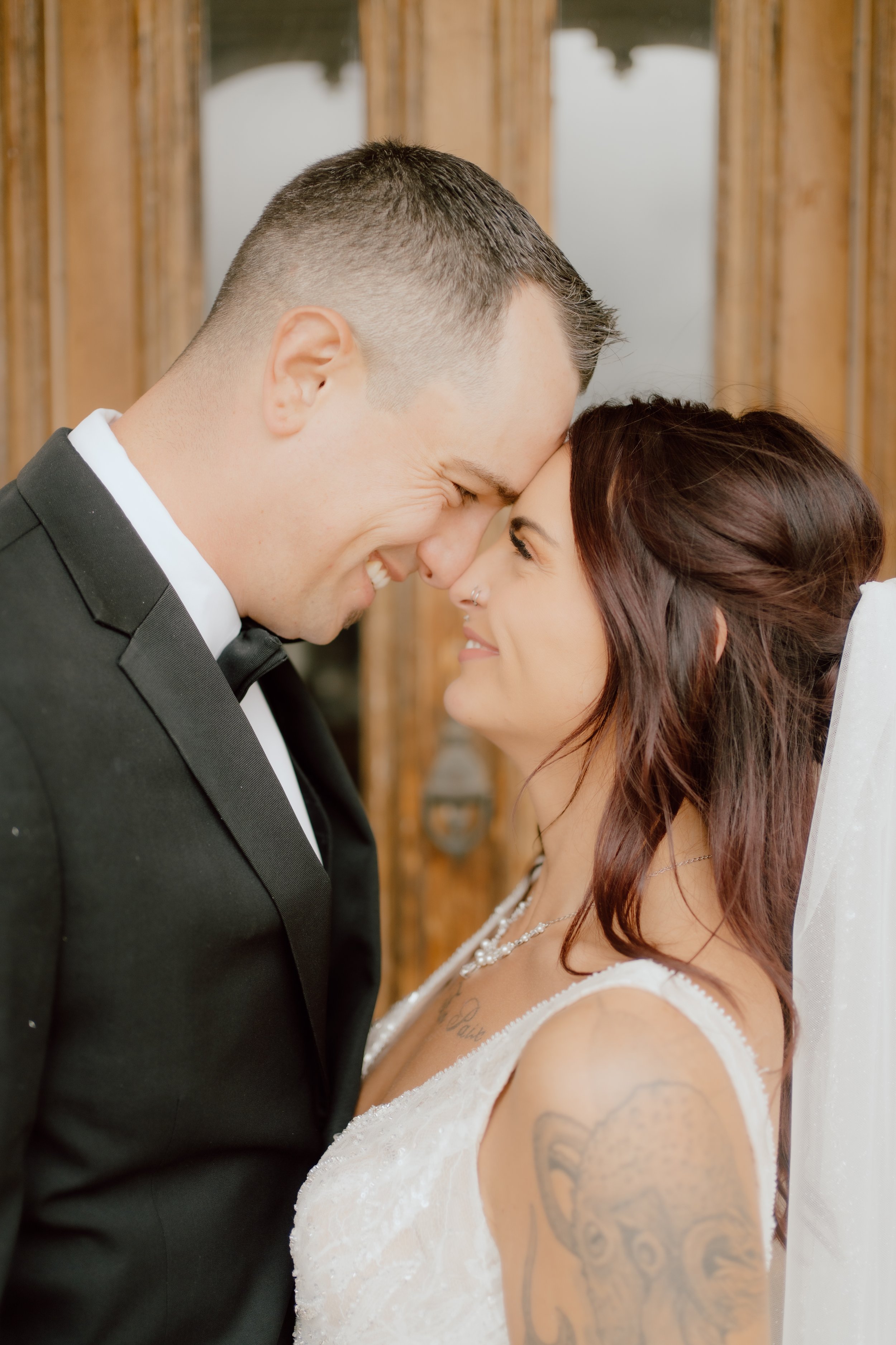 A couple, dressed in wedding attire, are touching foreheads and smiling at each other indoors with a wooden background.