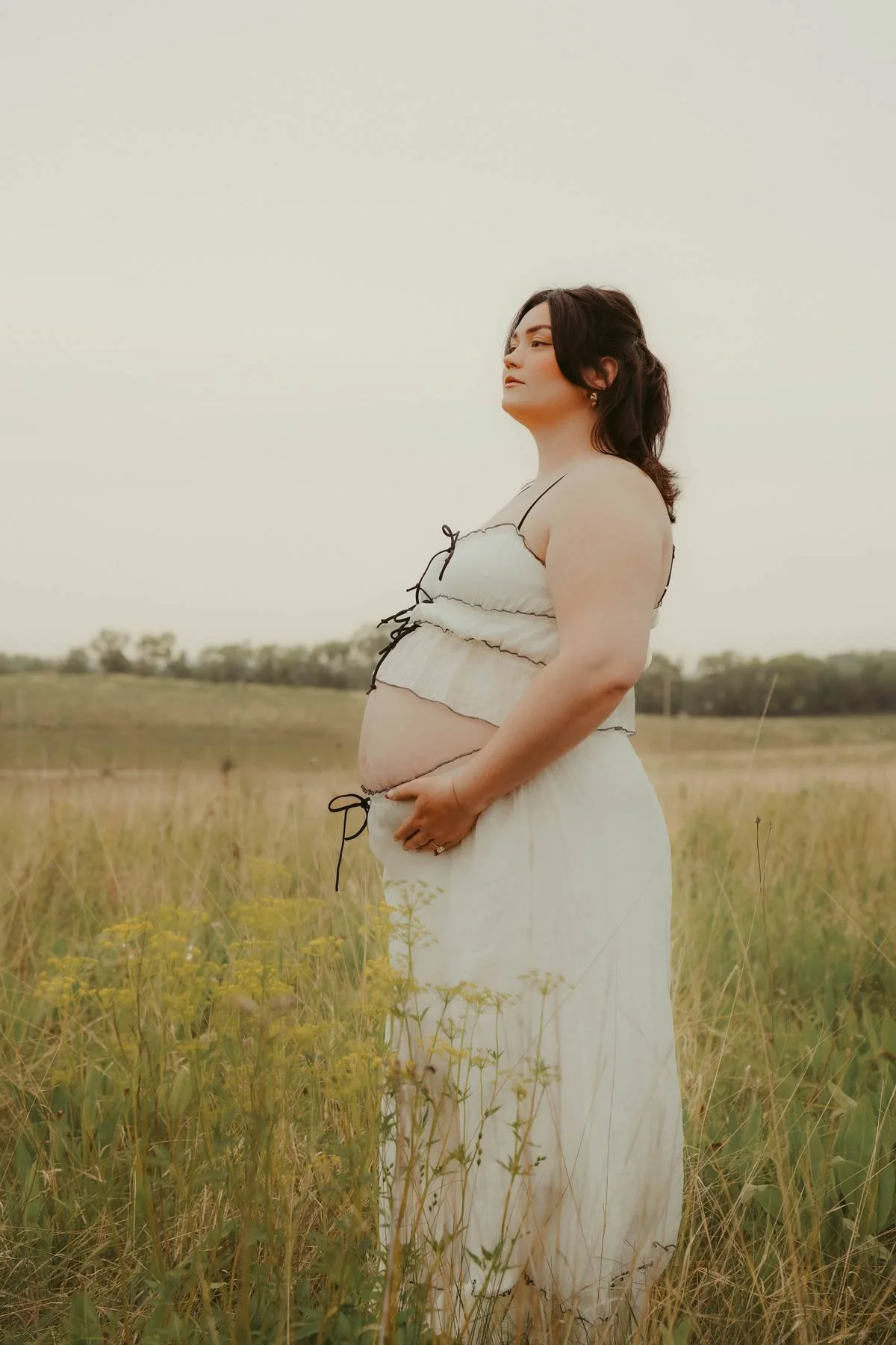 A pregnant woman standing in a grassy field during daytime, wearing a light-colored dress and looking to the side.