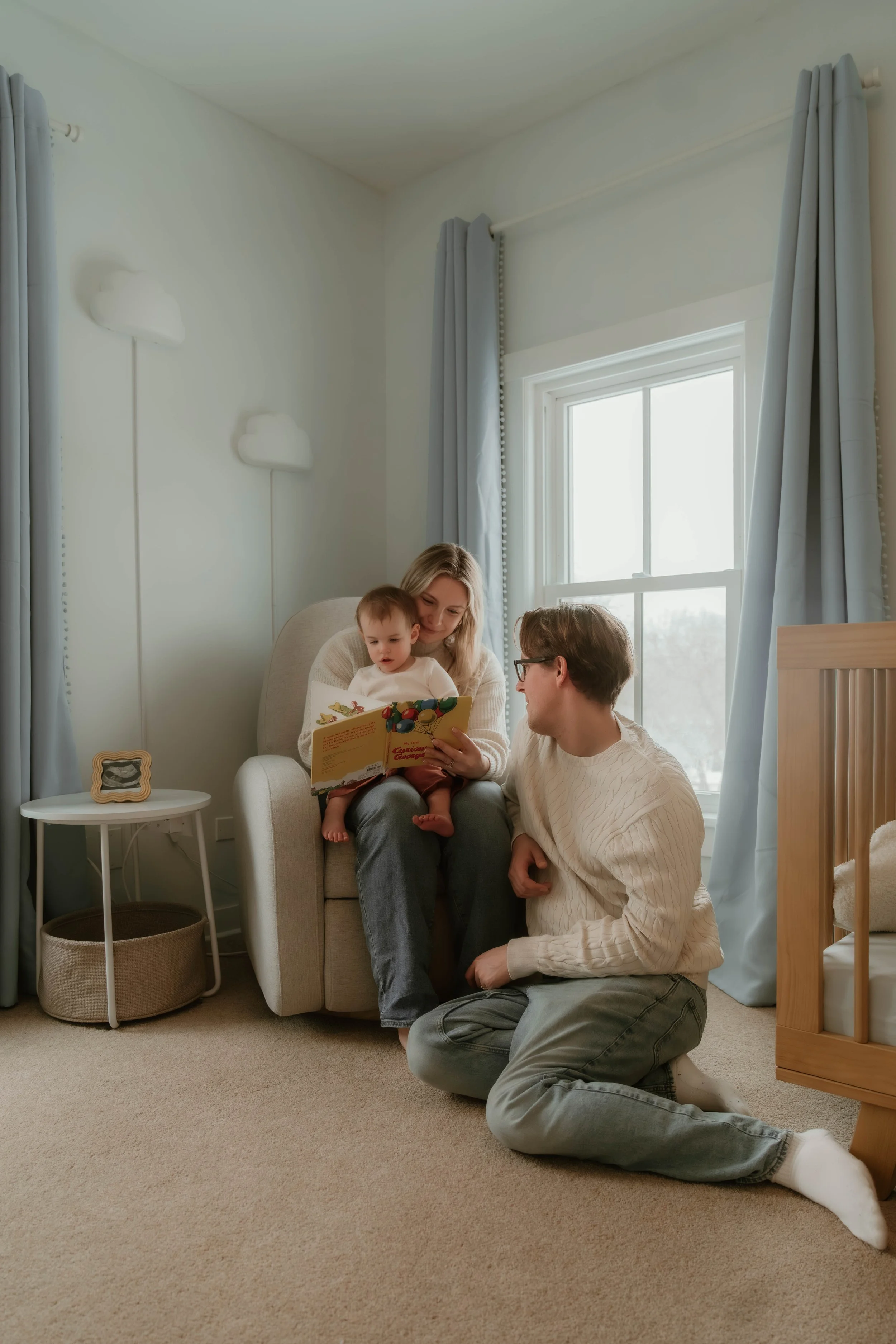 A family of three sitting in a cozy, softly lit nursery. The mother is sitting on a beige armchair holding a young child on her lap, showing him a colorful children's book. The father is sitting on the floor across from them, engaging with both. The 
