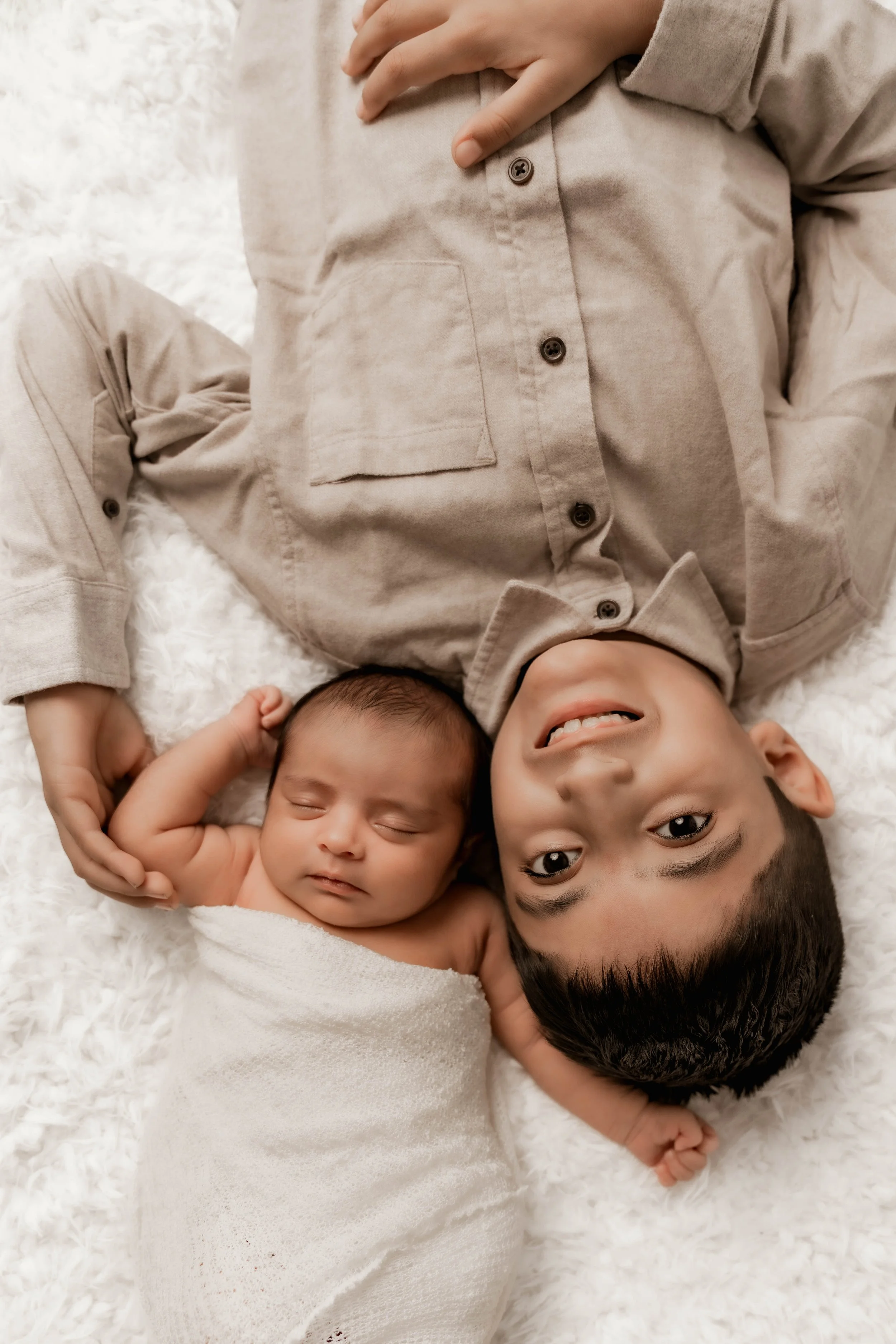A young boy lying on a fluffy white rug, smiling and looking up at the camera, with a newborn baby swaddled in white and sleeping beside him.