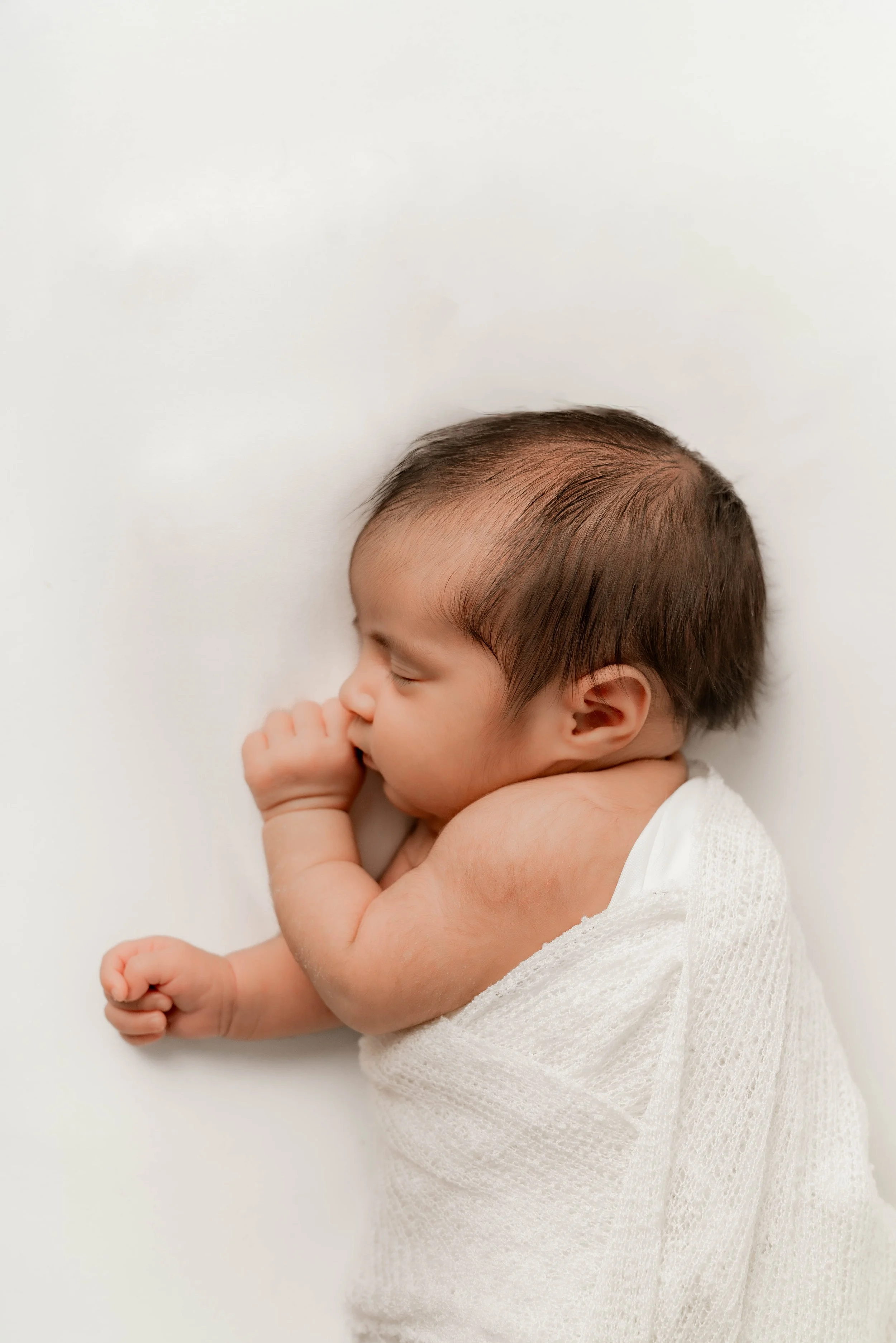 A sleeping baby wrapped in a white blanket, lying on a white surface, with hair tousled and eyes closed.