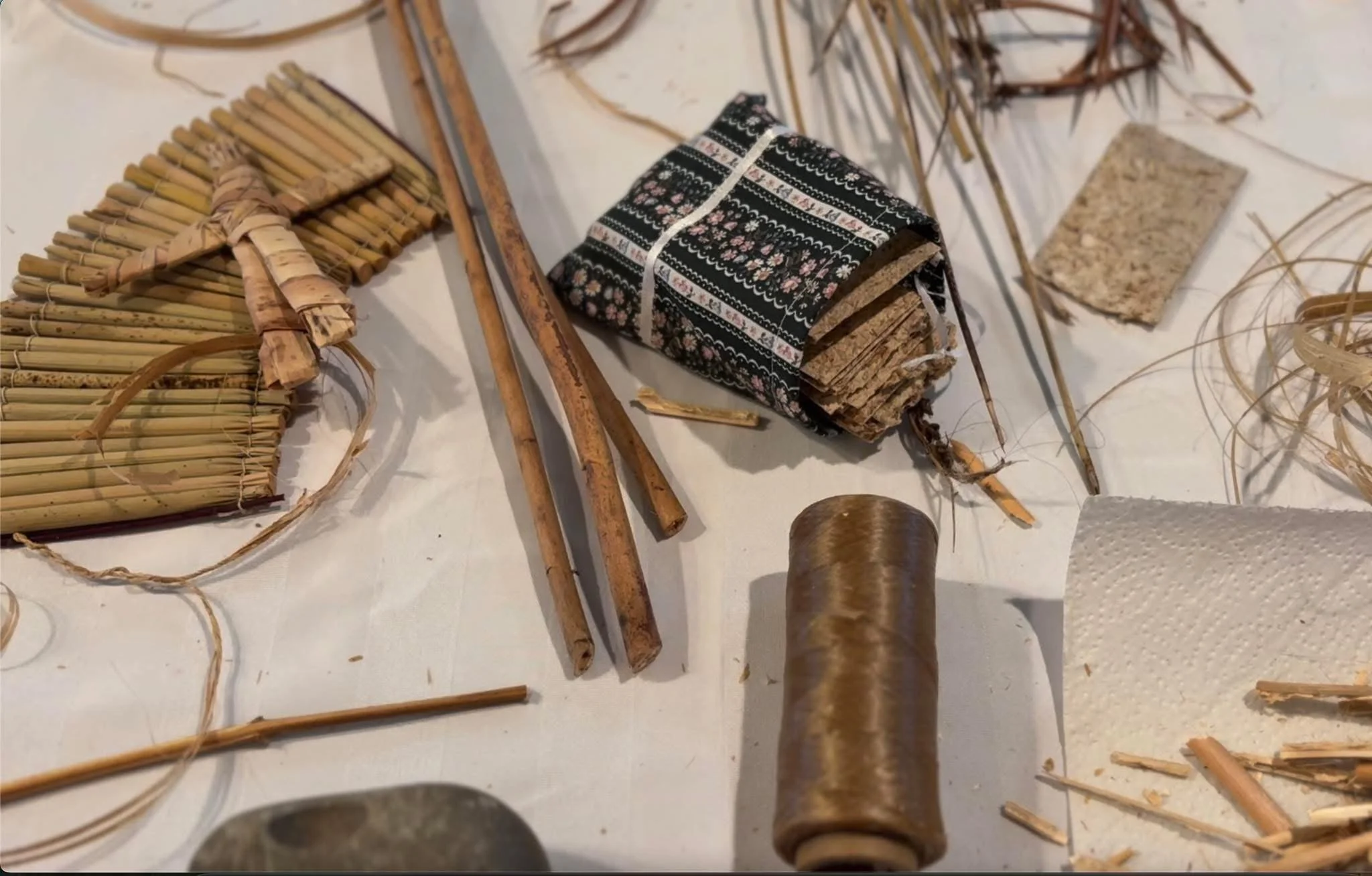 Collection of handmade natural craft materials, including bamboo sticks, a roll of woven fabric, a bamboo or wooden cylinder, dry grass or straw, a piece of fabric or paper, and a paper towel roll.