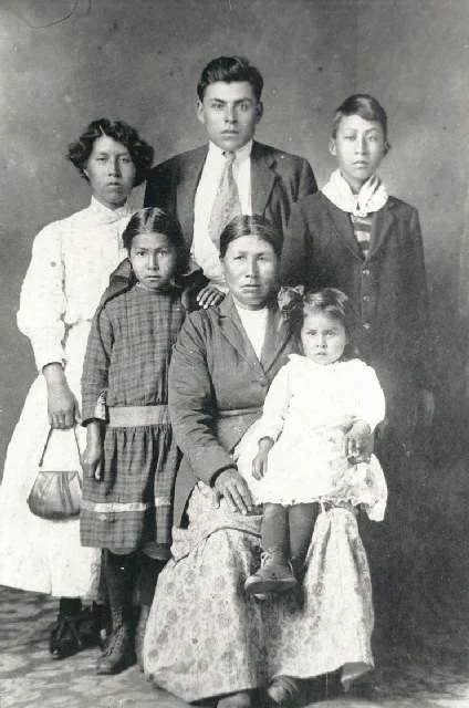 A black-and-white photograph of a family portrait with six people, including an adult woman seated with four children around her and one young girl sitting on her lap, all dressed in vintage clothing.