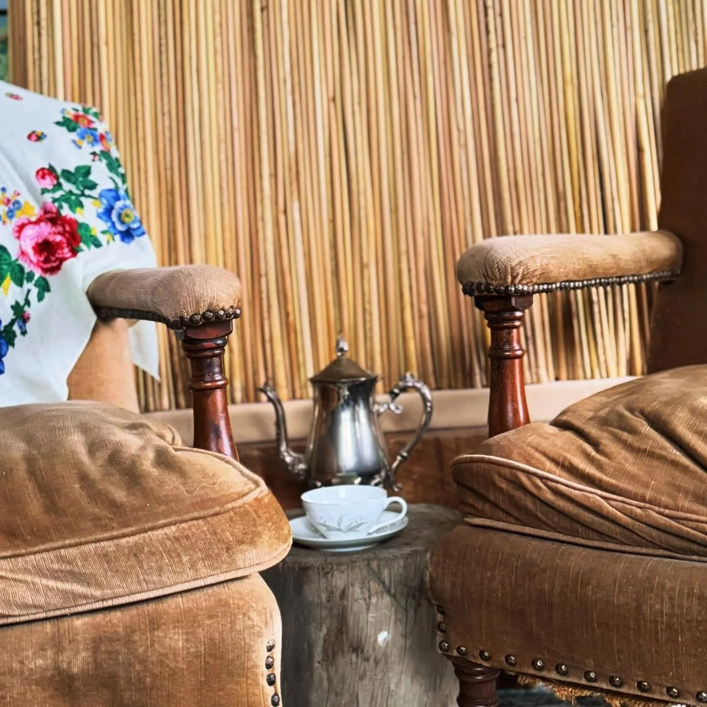 Vintage interior with velvet armchairs, a metal teapot, a teacup, and saucer on a small wooden table against a background of bamboo wall paneling and a floral embroidered pillow.